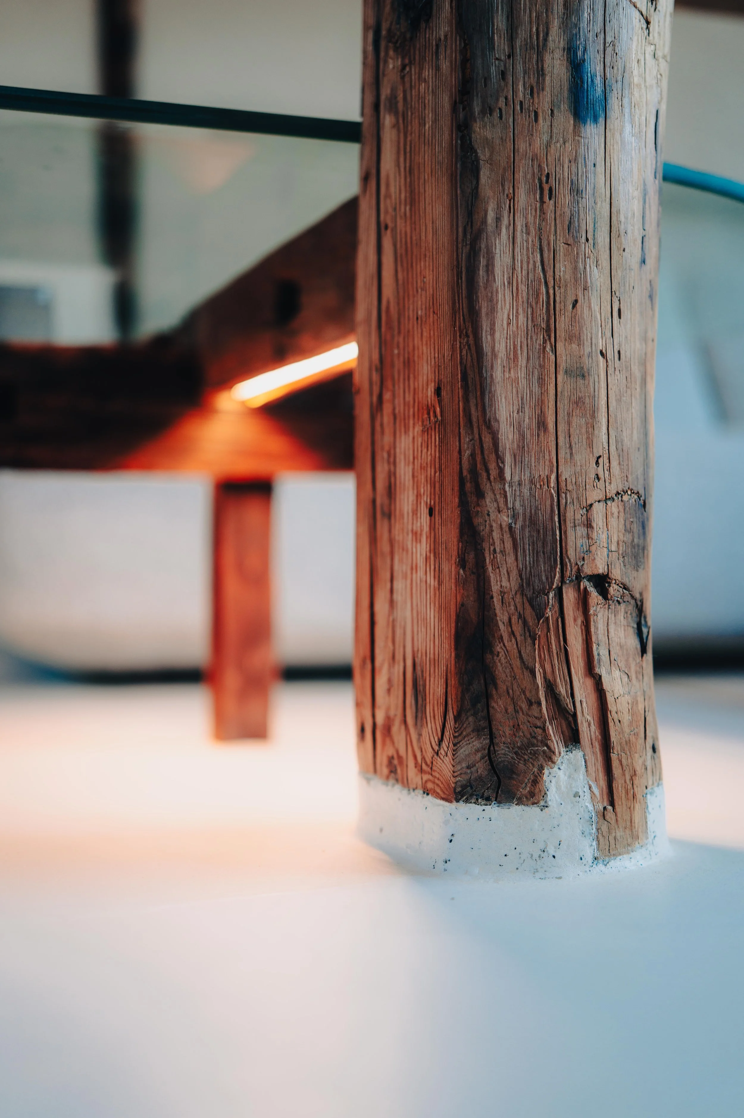 NUUT Aewol 눗애월 Close-up of a wooden support post with visible cracks and natural texture, anchored to a white base, with blurred background.