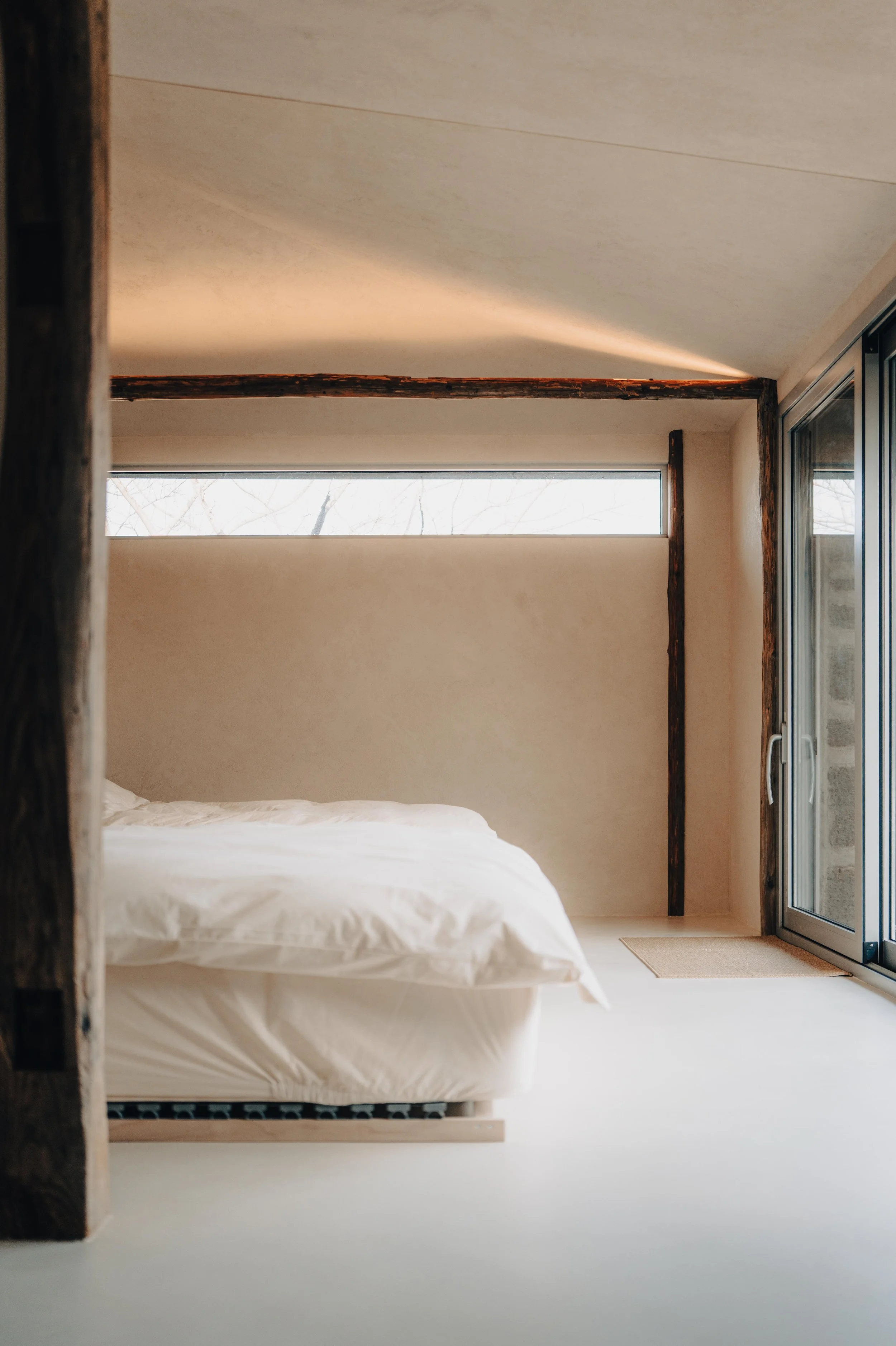 NUUT Aewol 눗애월 Minimalist bedroom with a bed, wooden beams, a horizontal window, and a sliding glass door.