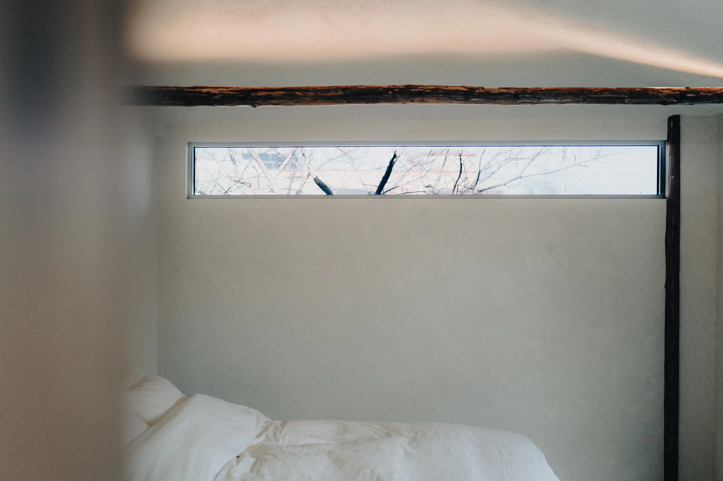 NUUT Aewol 눗애월 Partially visible bed with white bedding in a room with minimalist decor, featuring a horizontal window showing a snowy landscape with trees outside.