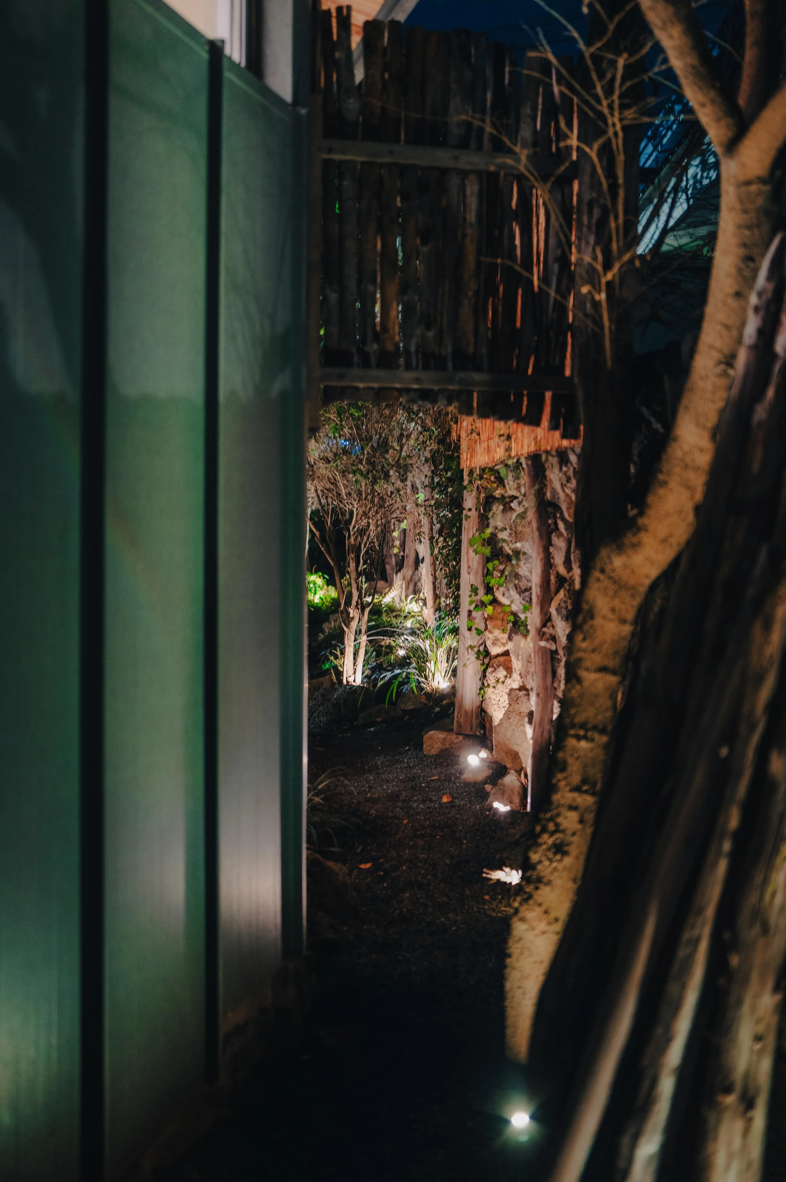 NUUT Aewol 눗애월 Nighttime garden pathway illuminated by ground lights, surrounded by trees and wooden fence.