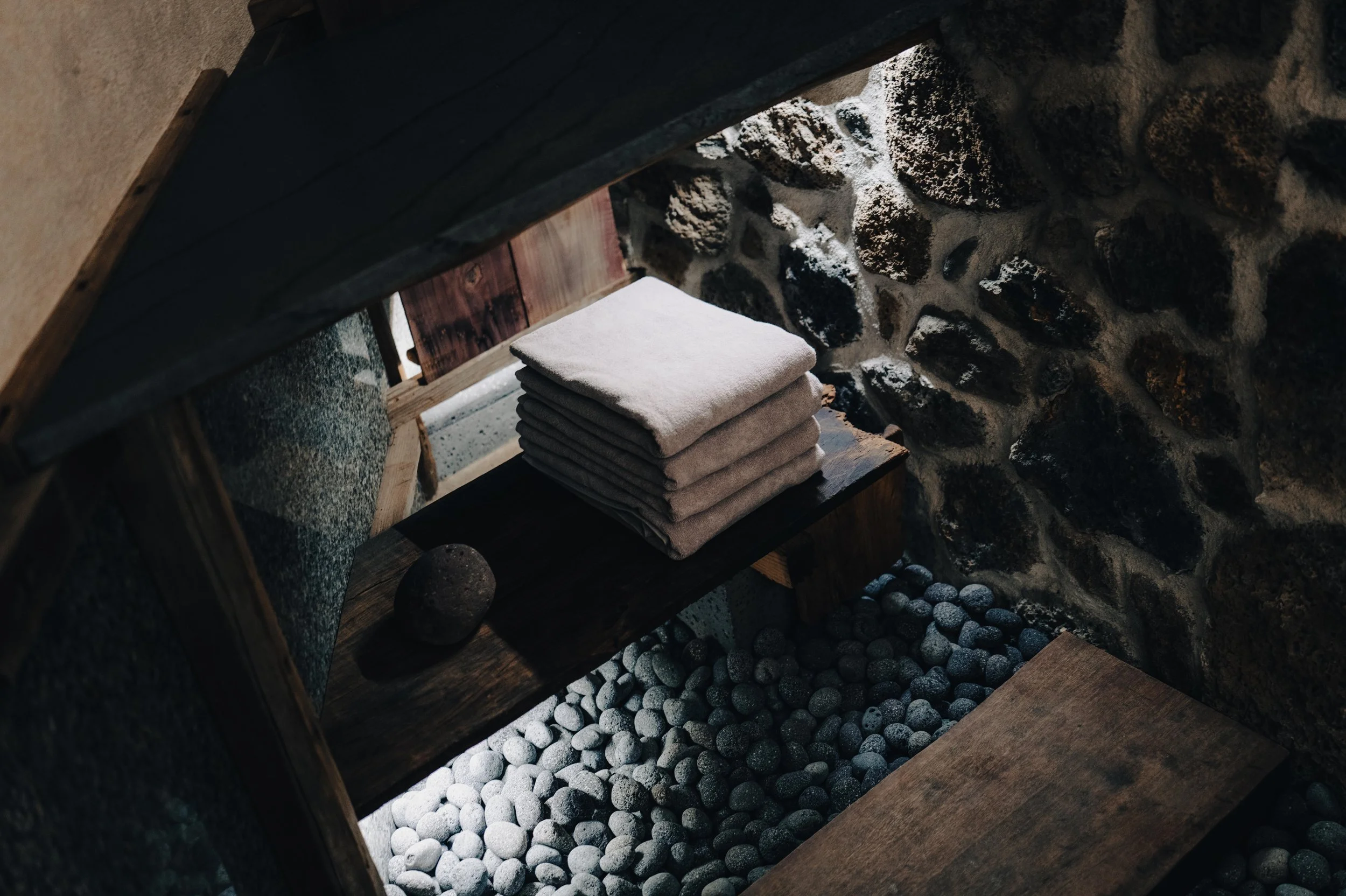 NUUT Aewol 눗애월 A stack of six folded towels on a wooden shelf inside a rustic stone-walled room, with stones on the floor and a stone wall background.