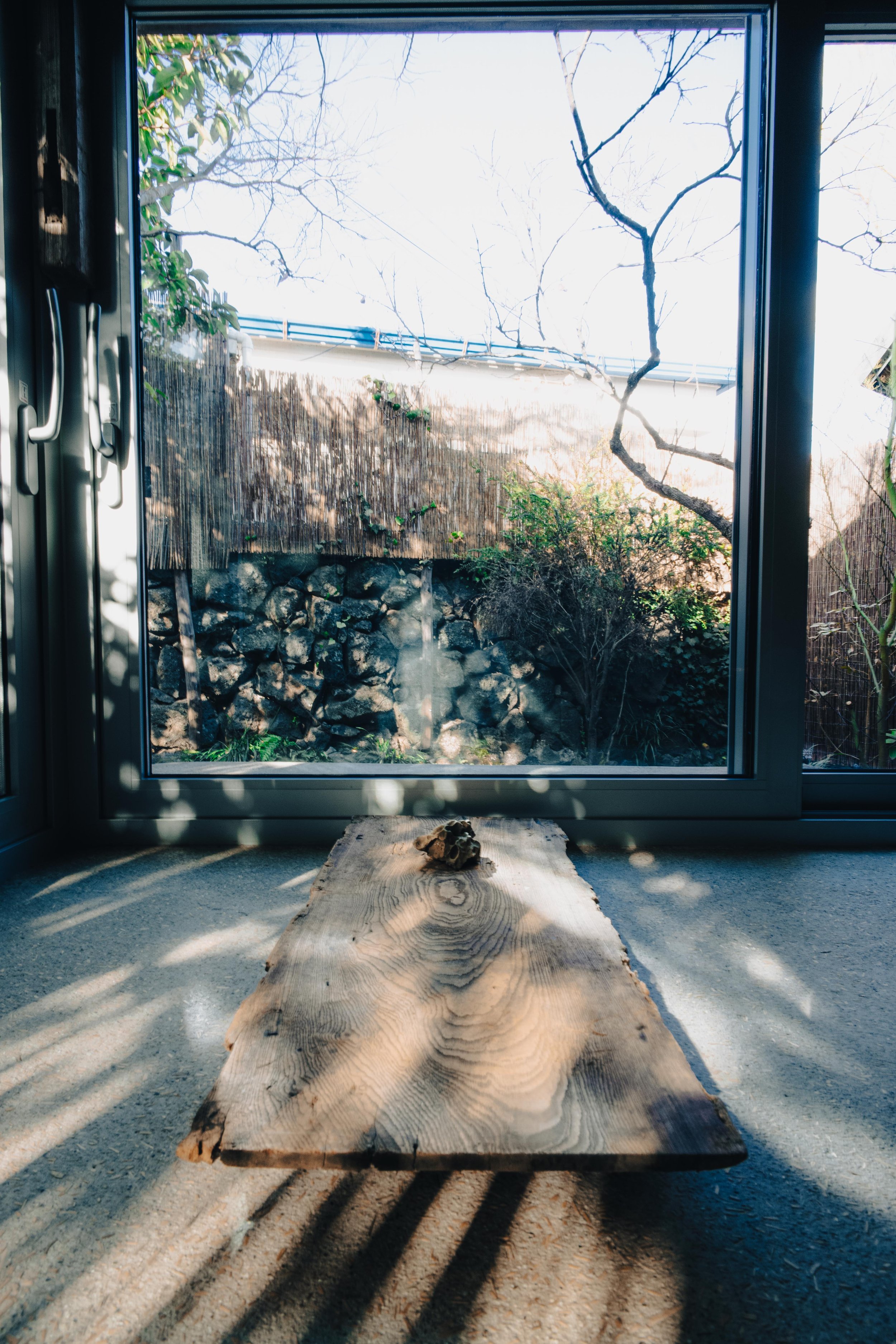 Indoor scene with a large glass door showing an outdoor garden with bare trees, rocks, plants, and a bamboo fence. Inside, a wooden slab with a small rock on top rests on a concrete floor, with sunlight casting shadows through the glass.
