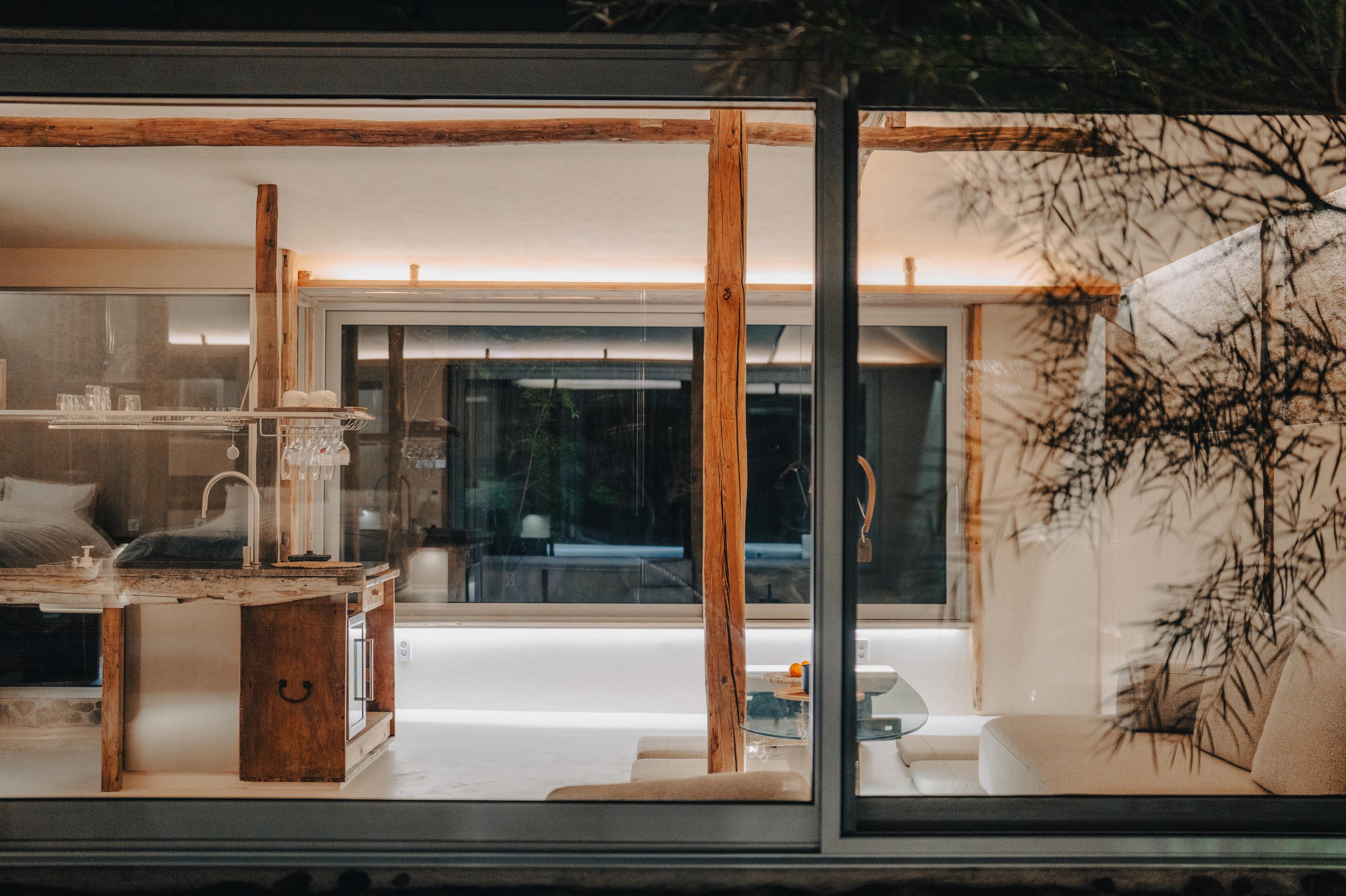 NUUT Aewol 눗애월 Interior view of a modern, minimalist living space seen through large glass windows, featuring a dining area with a small round table, a sofa with cushions, and wooden structural elements.