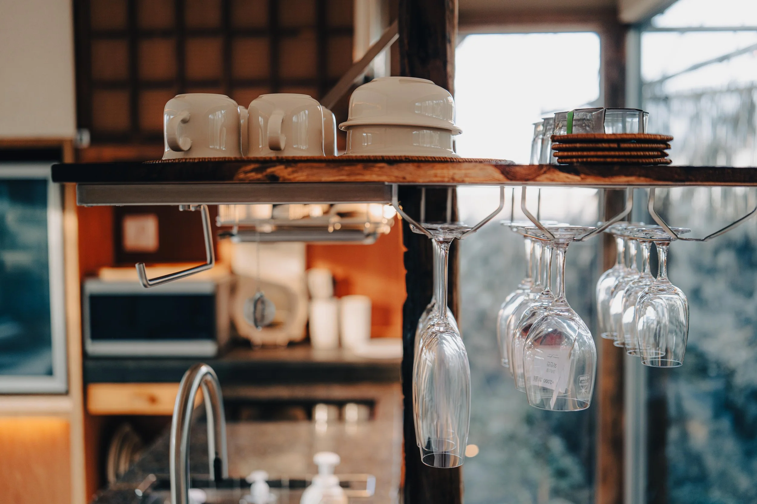 NUUT Aewol 눗애월 A wooden shelf holding white cups, stacked plates, and hanging wine glasses in a cozy kitchen with natural light.