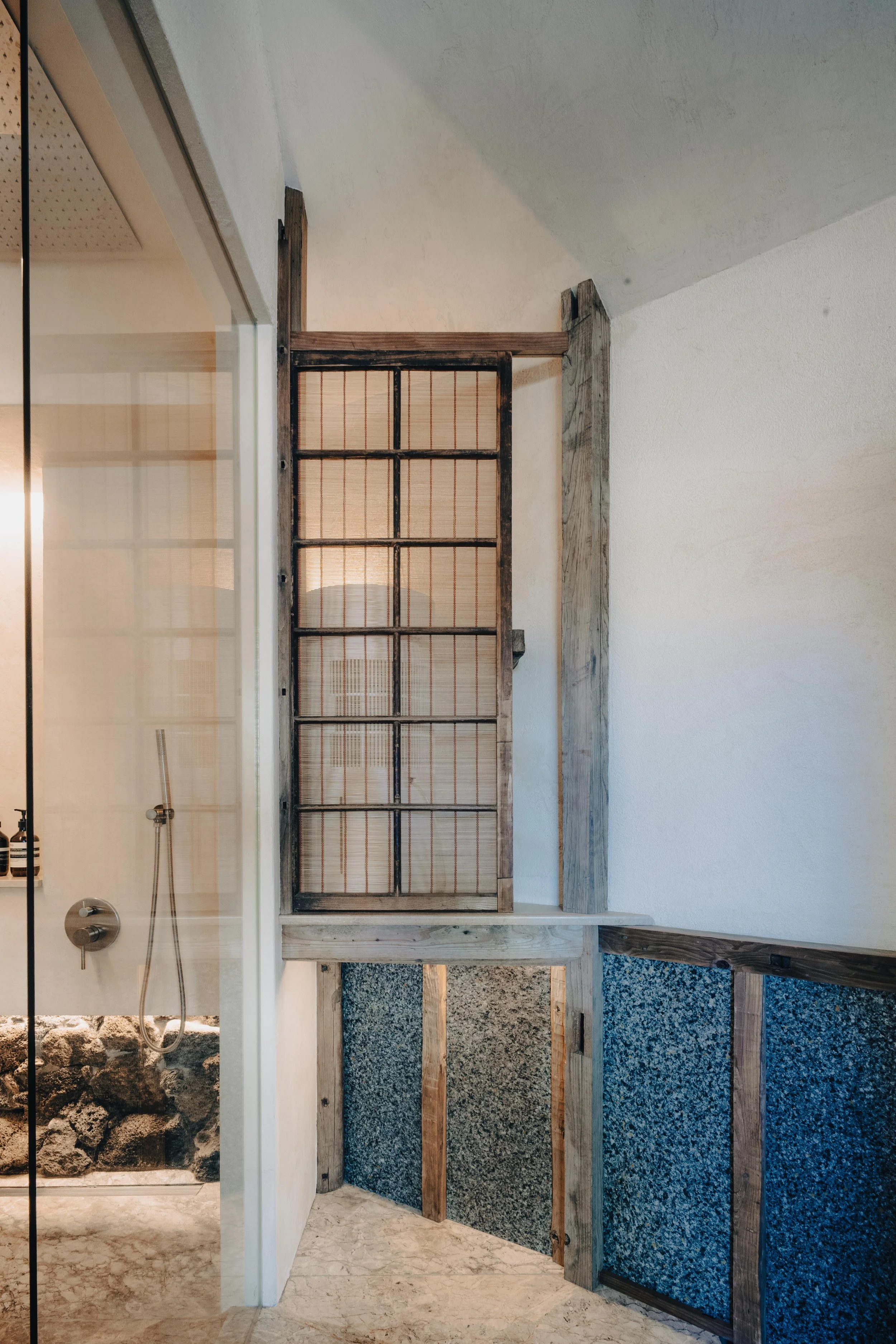 NUUT Aewol 눗애월 Interior corner of a room with a shower area on the left and rustic wood framing and panels, some with stone and textured surfaces, on the right.