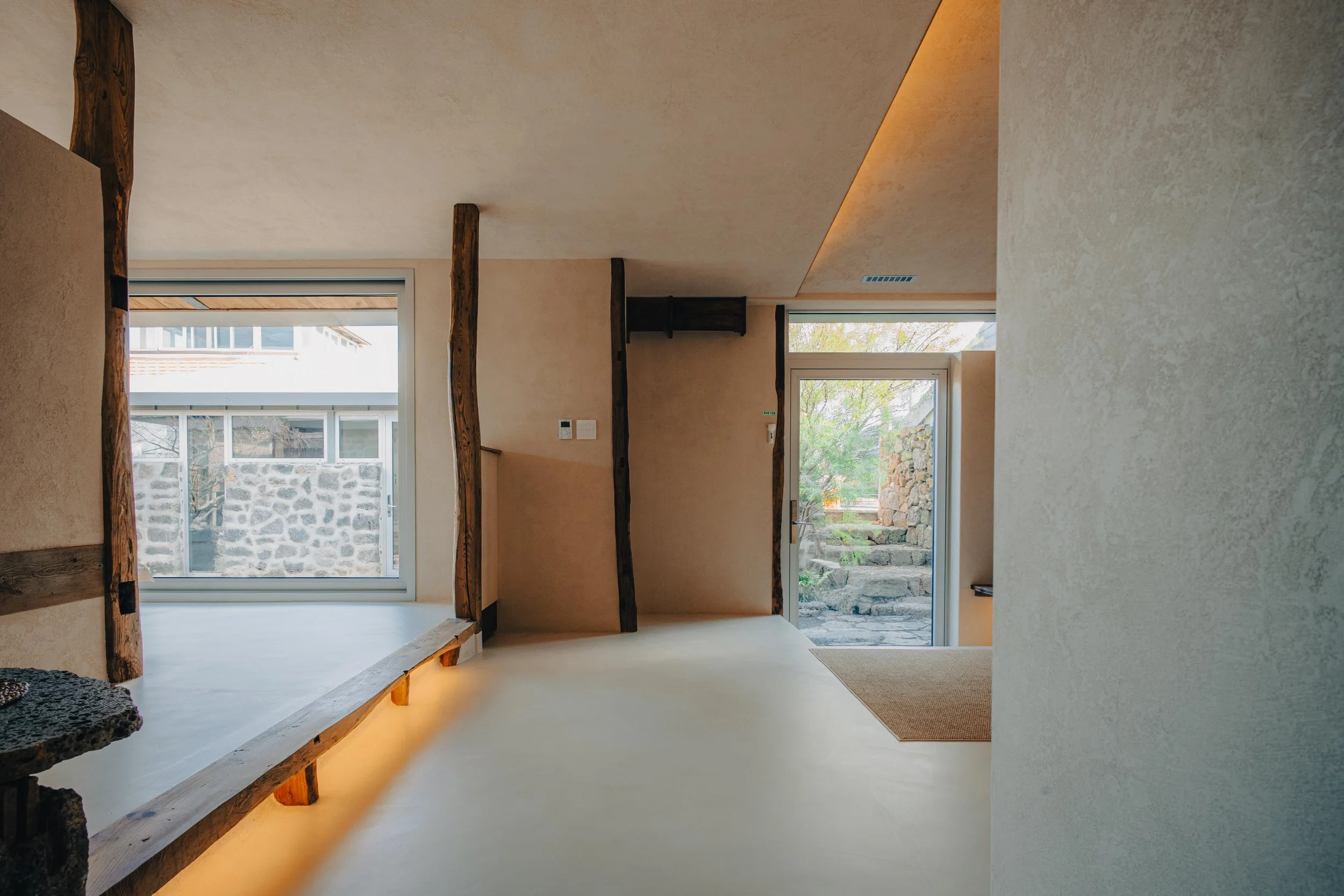 NUUT Aewol 눗애월 Interior of a modern house with textured beige walls, dark wooden beams, a large window, and a glass door leading outside to stone steps.