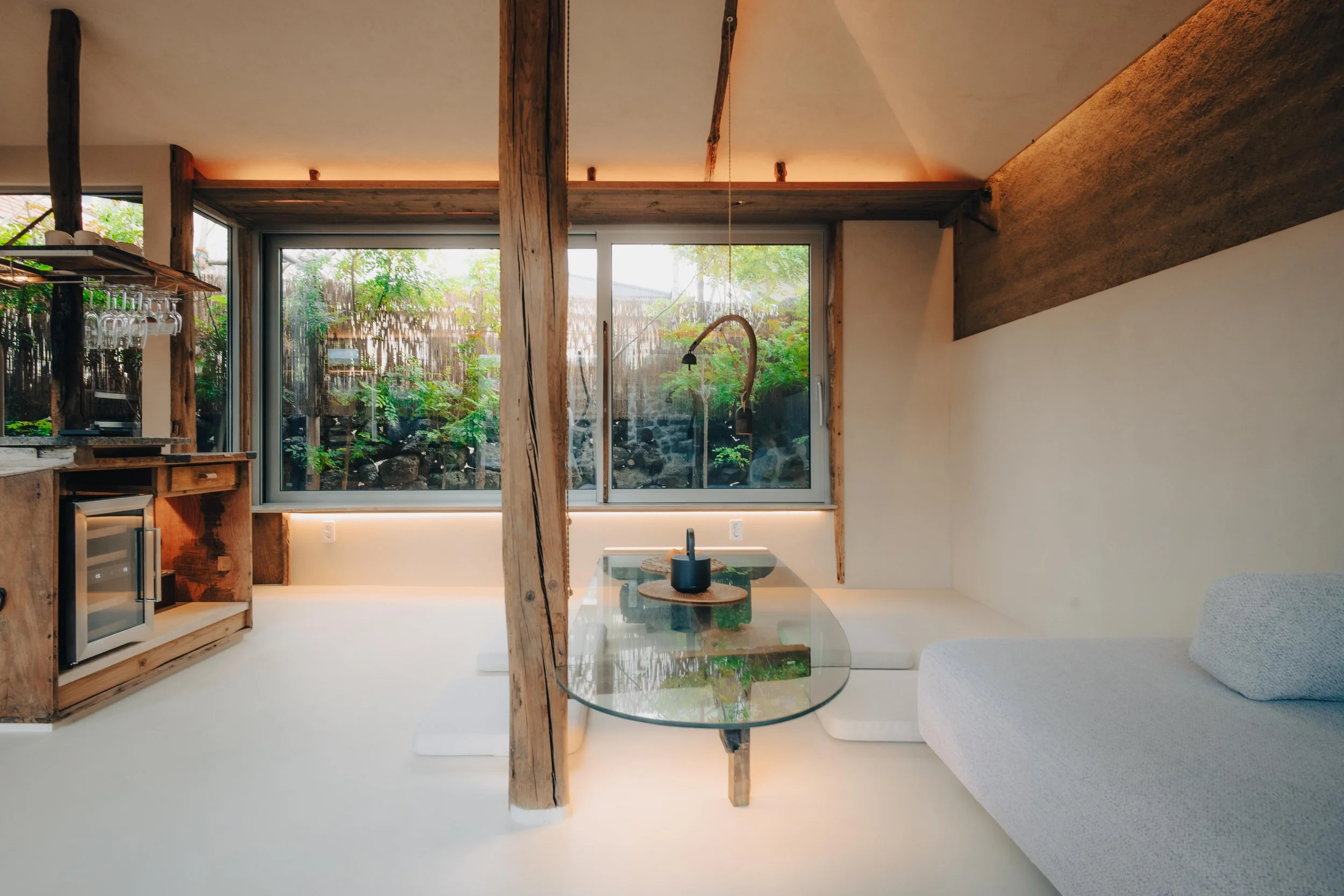 NUUT Aewol 눗애월 Interior of a modern, minimalist room with a glass table, a white cushioned seating area, wooden accents, and large windows overlooking a garden.