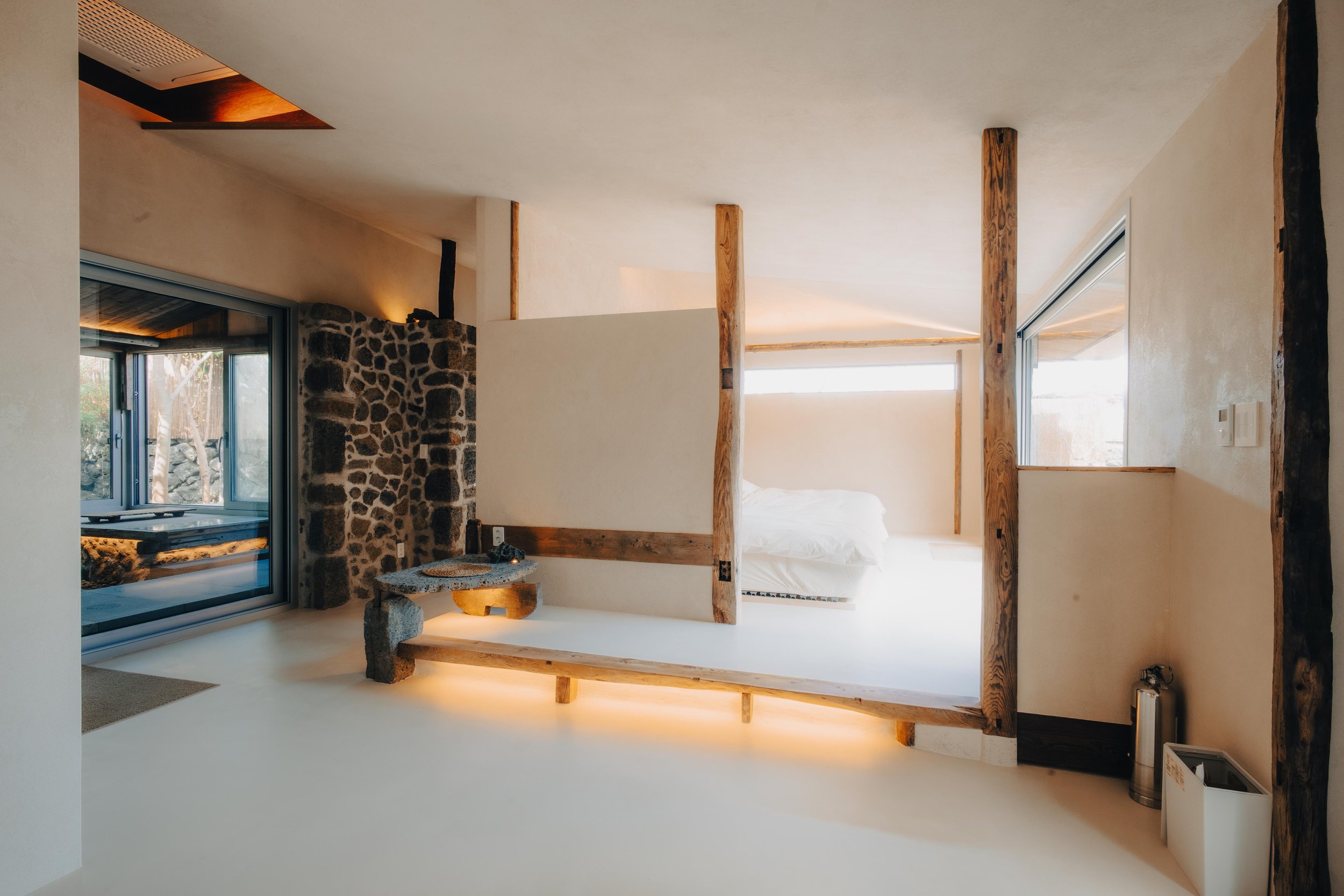 NUUT Aewol 눗애월 Modern bedroom with minimalist design, featuring a bed, wooden beams, stone wall, large window, and sliding glass door leading to an outdoor area.
