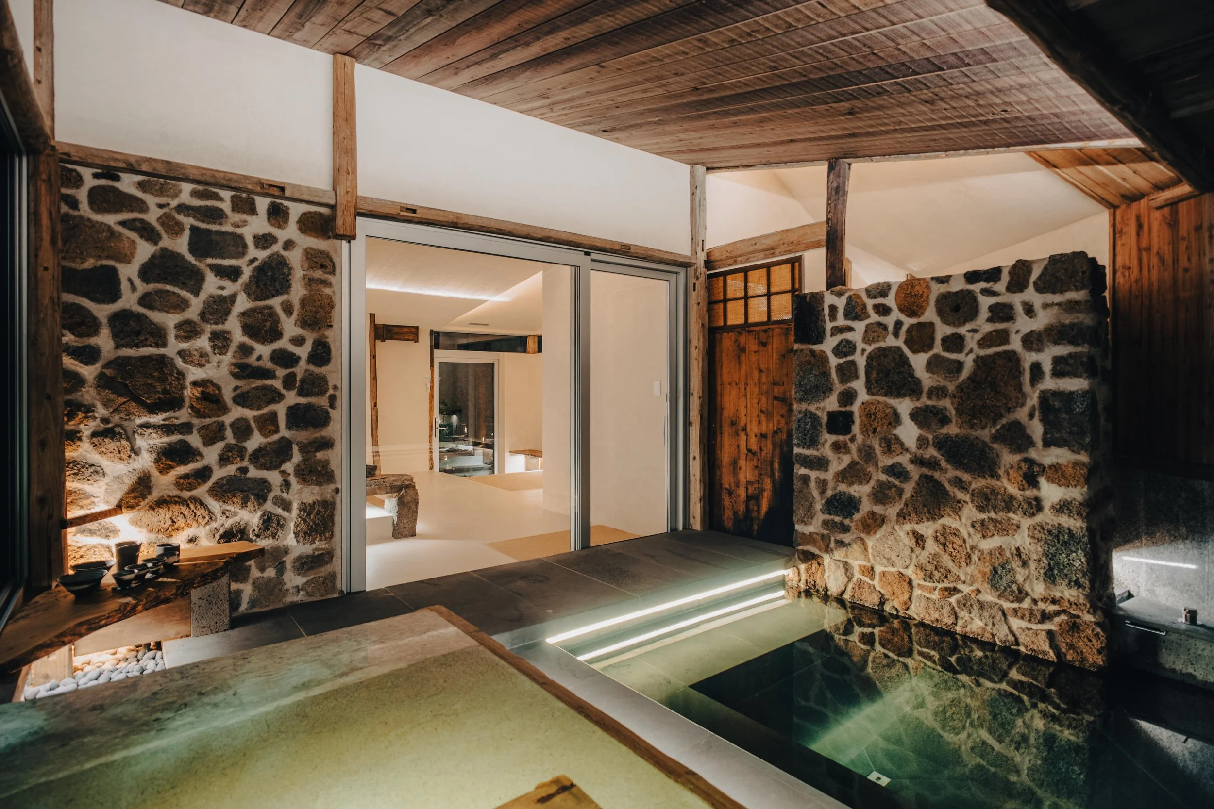 NUUT Aewol 눗애월 Indoor space with stone and wood walls, featuring a hot tub with water, illuminated steps, and sliding glass doors leading to another room.