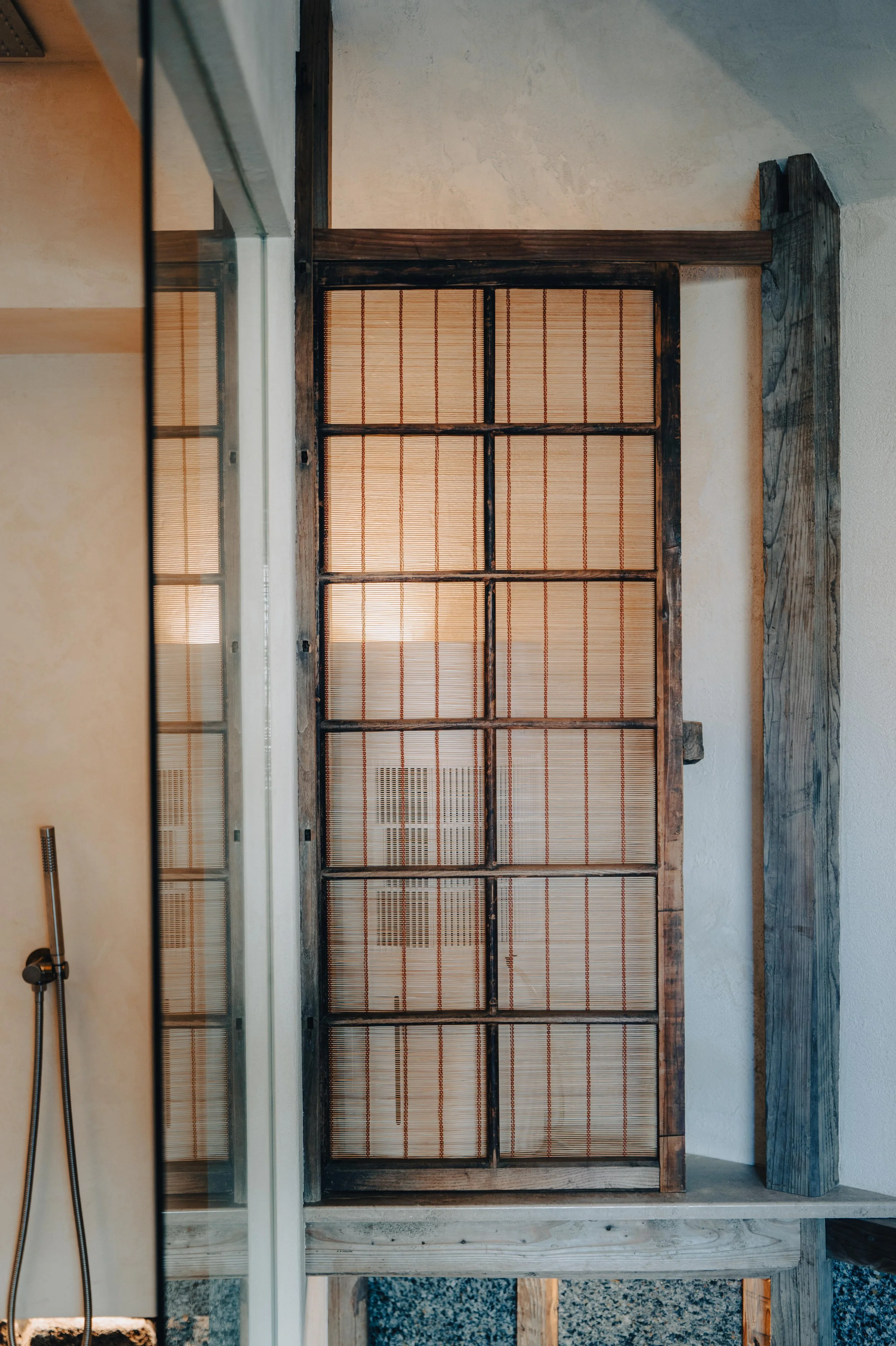 NUUT Aewol 눗애월 A wooden frame with a bamboo or straw screen, part of a room or outdoor structure, with a corner of a shower or similar fixture visible on the left side.