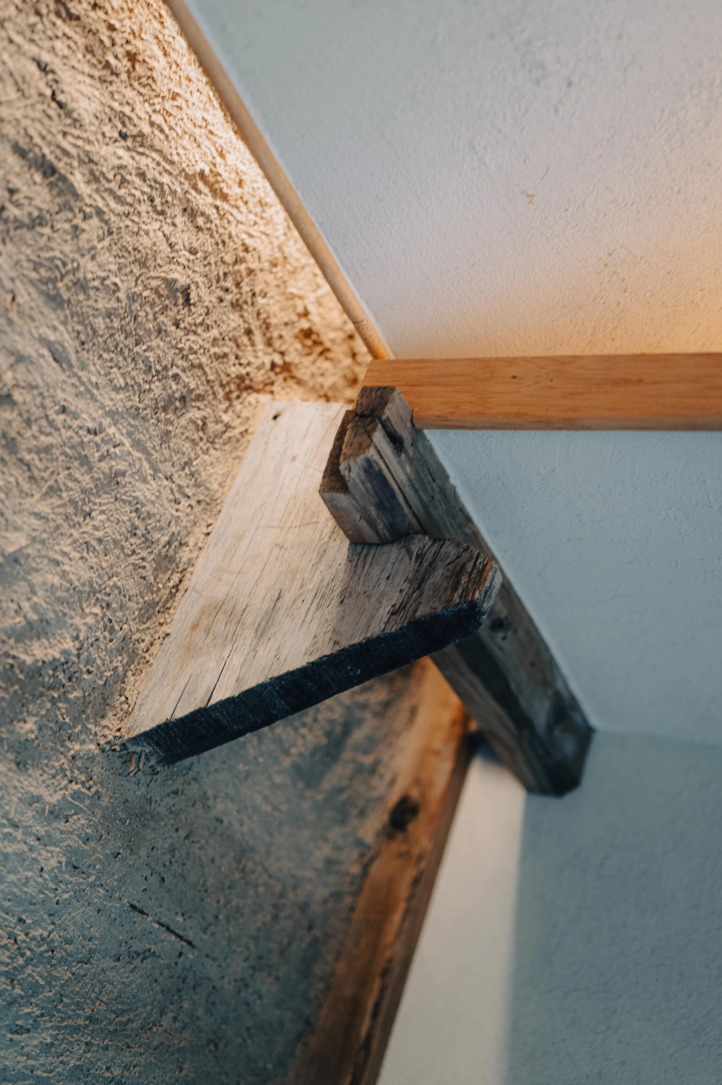 NUUT Aewol 눗애월 Close-up of a wooden shelf supported by a small wood block, with drywall and a textured wall in the background.