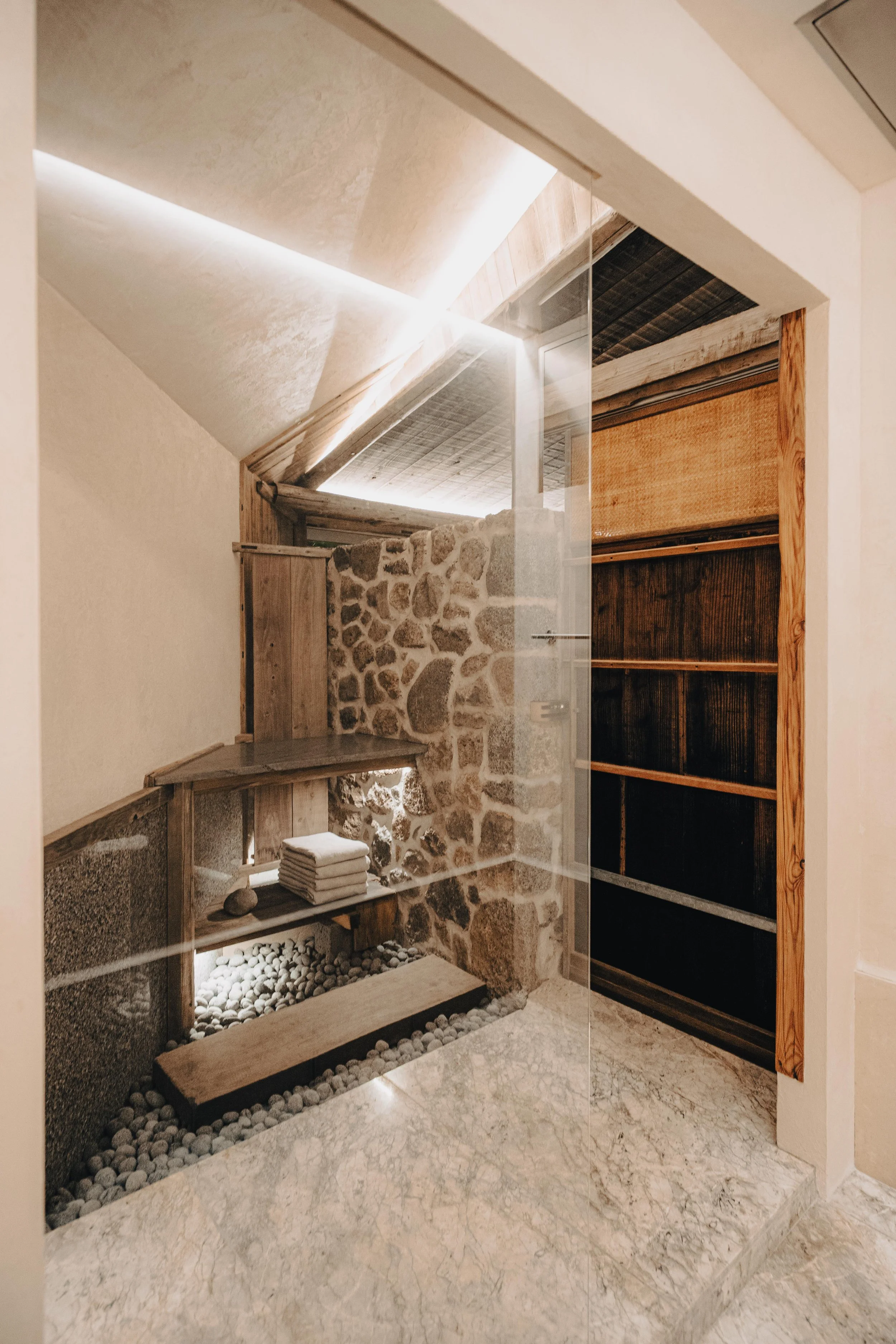 NUUT Aewol 눗애월 Interior of a spa room with stone and wooden accents, a small bench, and shelves, viewed through a glass door.