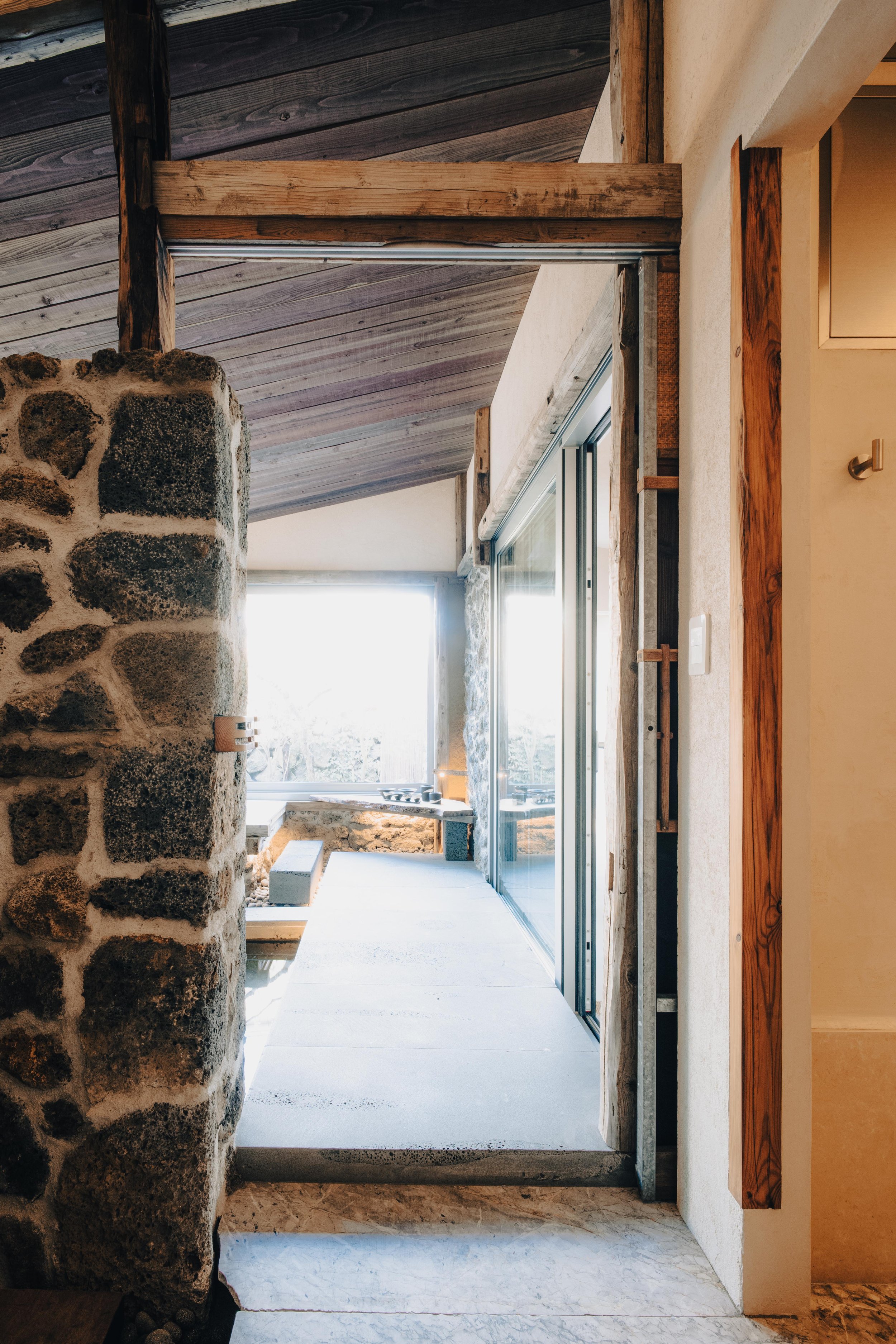 NUUT Aewol 눗애월 Interior view of a room with a stone wall, wooden ceiling, and sliding glass door leading to an outdoor space with snow-covered ground.