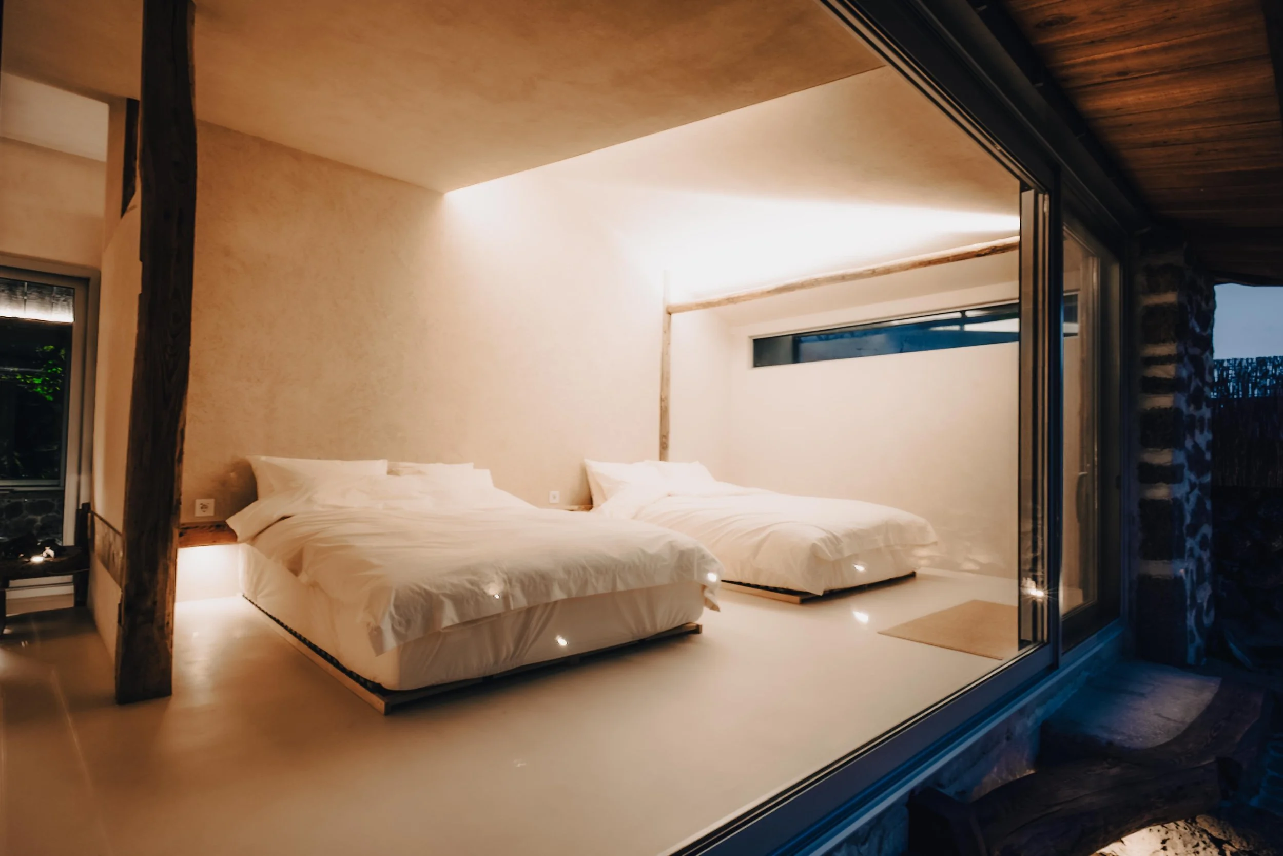 NUUT Aewol 눗애월 Modern bedroom with two white beds and minimalist decor, enclosed with glass walls.