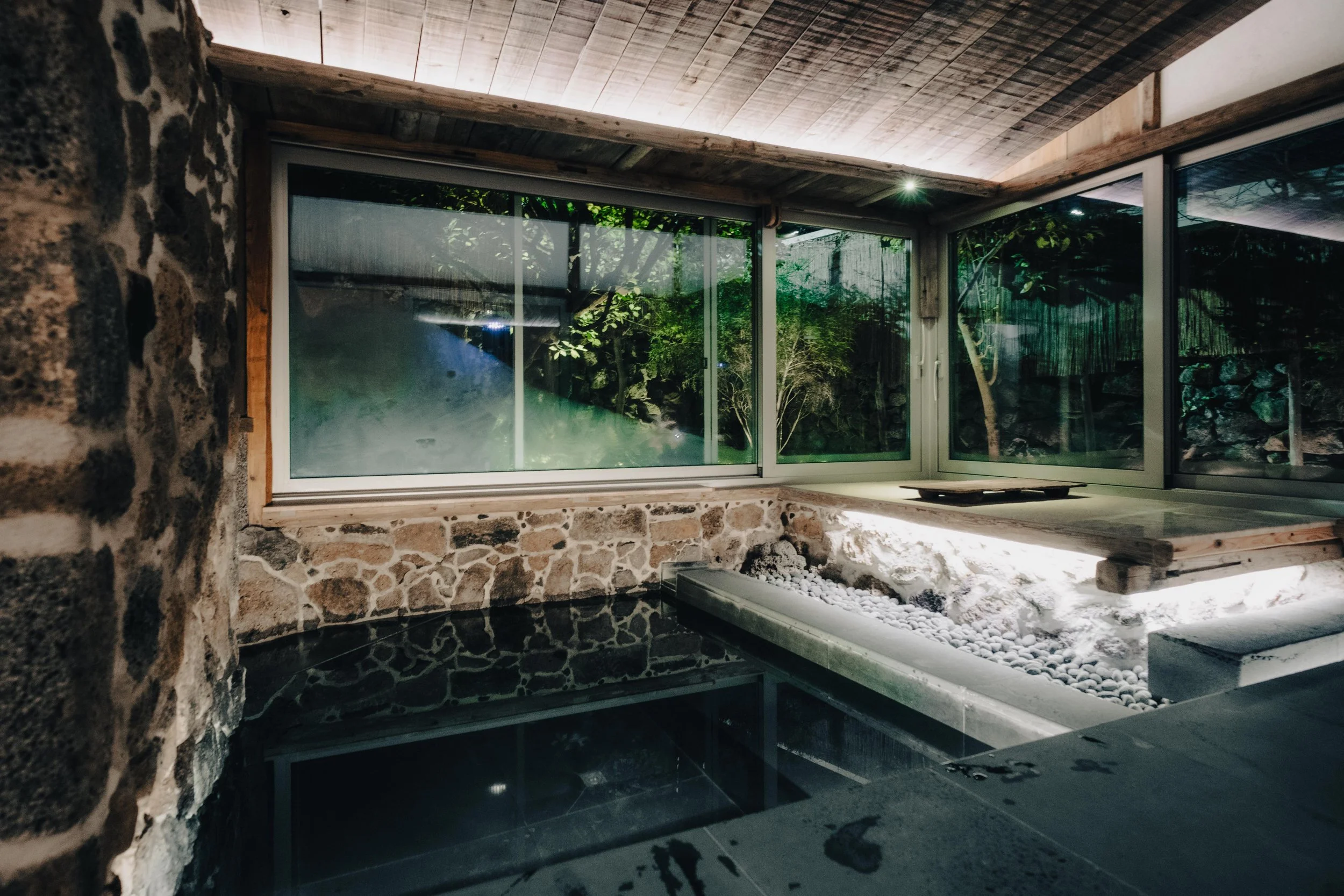 NUUT Aewol 눗애월 Indoor spa with stone walls, large glass windows showing a garden, and rocks with water features inside.