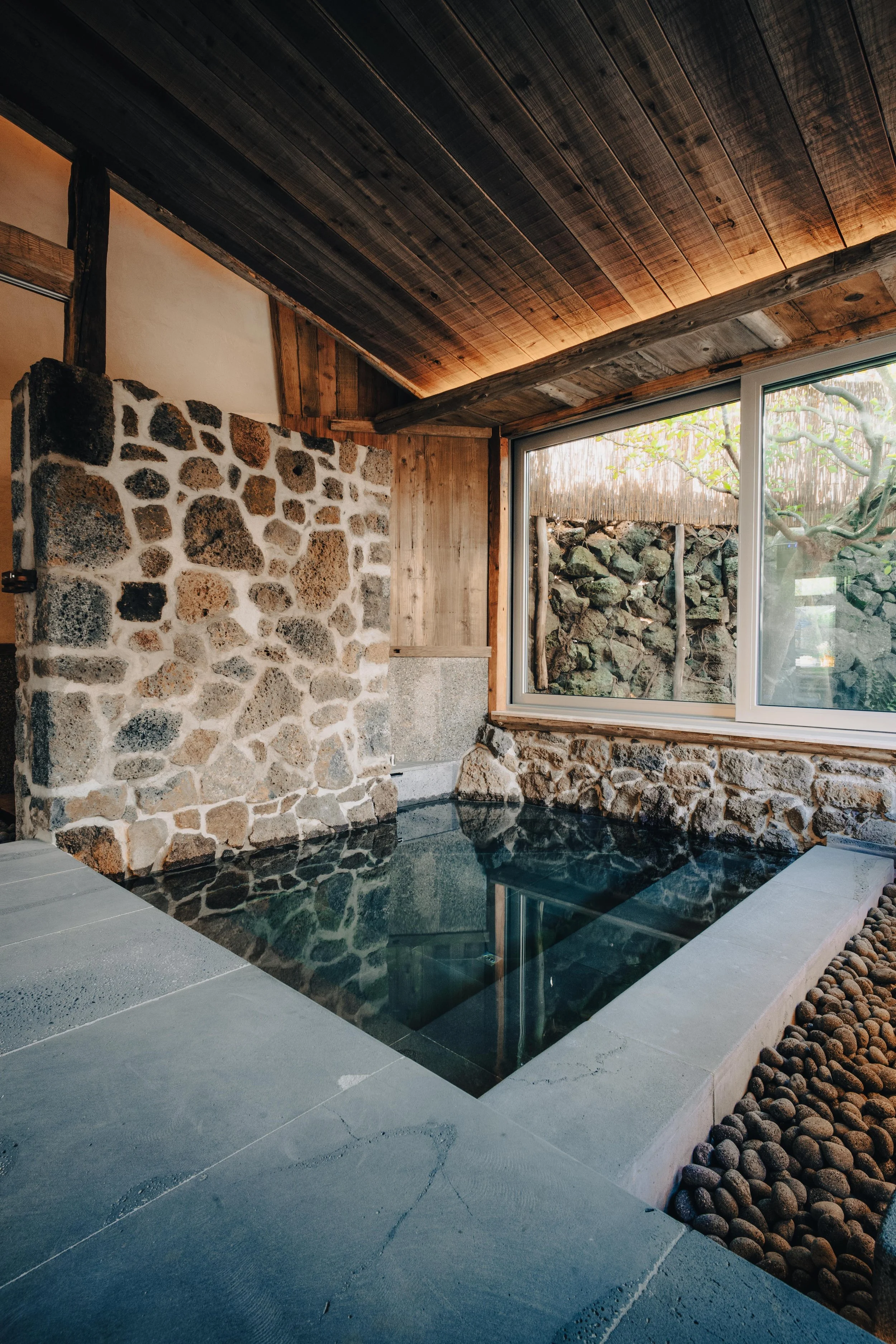 Indoor Japanese-style stone bath with a stone wall, large window overlooking a rocky garden, and a wooden ceiling.