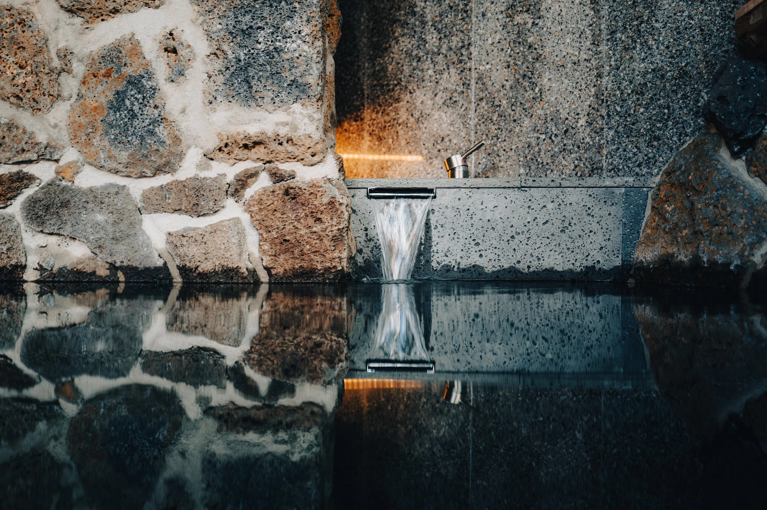 Indoor fountain with a water stream flowing from a rectangular stone basin, against a backdrop of rustic stone and gray textured wall.