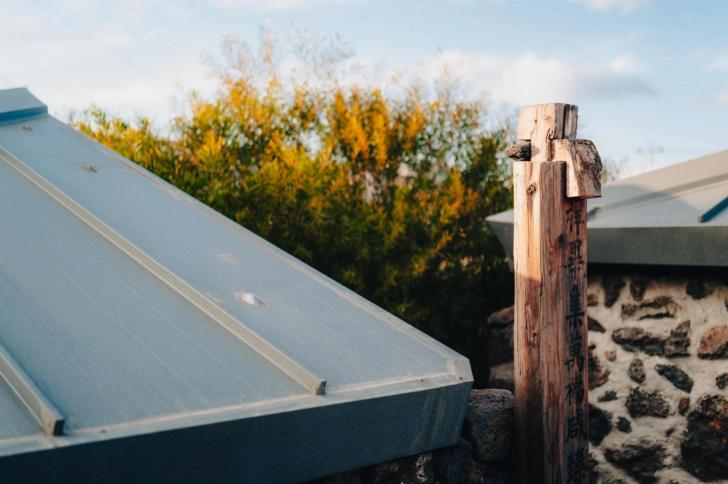 NUUT Aewol 눗애월 Close-up of a solar panel on a roof, with a weathered wooden post with Japanese writing next to it. In the background, there are trees with autumn-colored leaves and part of another roof. The scene is outdoors during daytime.
