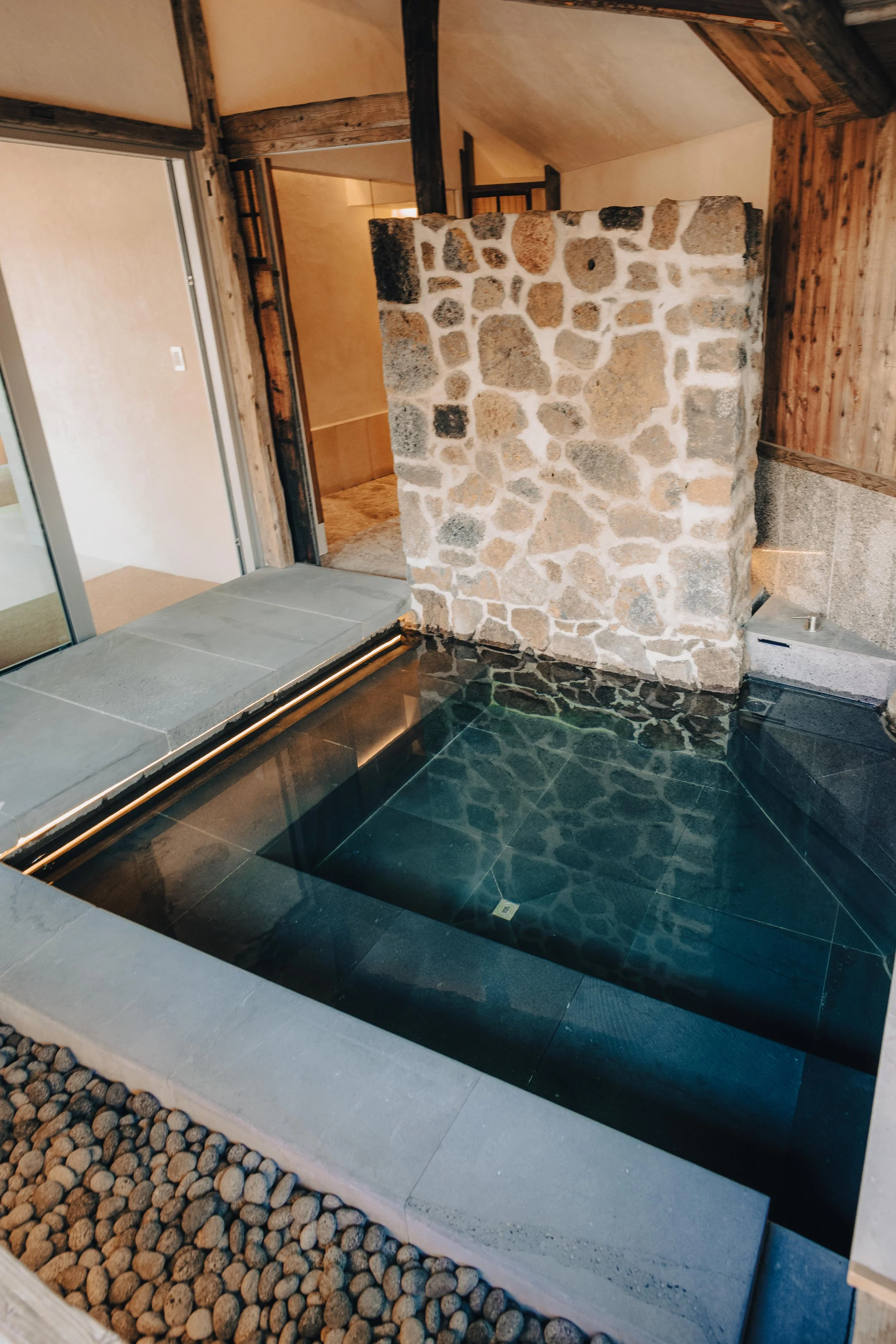 NUUT Aewol 눗애월 Indoor hot tub with dark tile, stone wall, wooden accents, and a glass door leading outside.