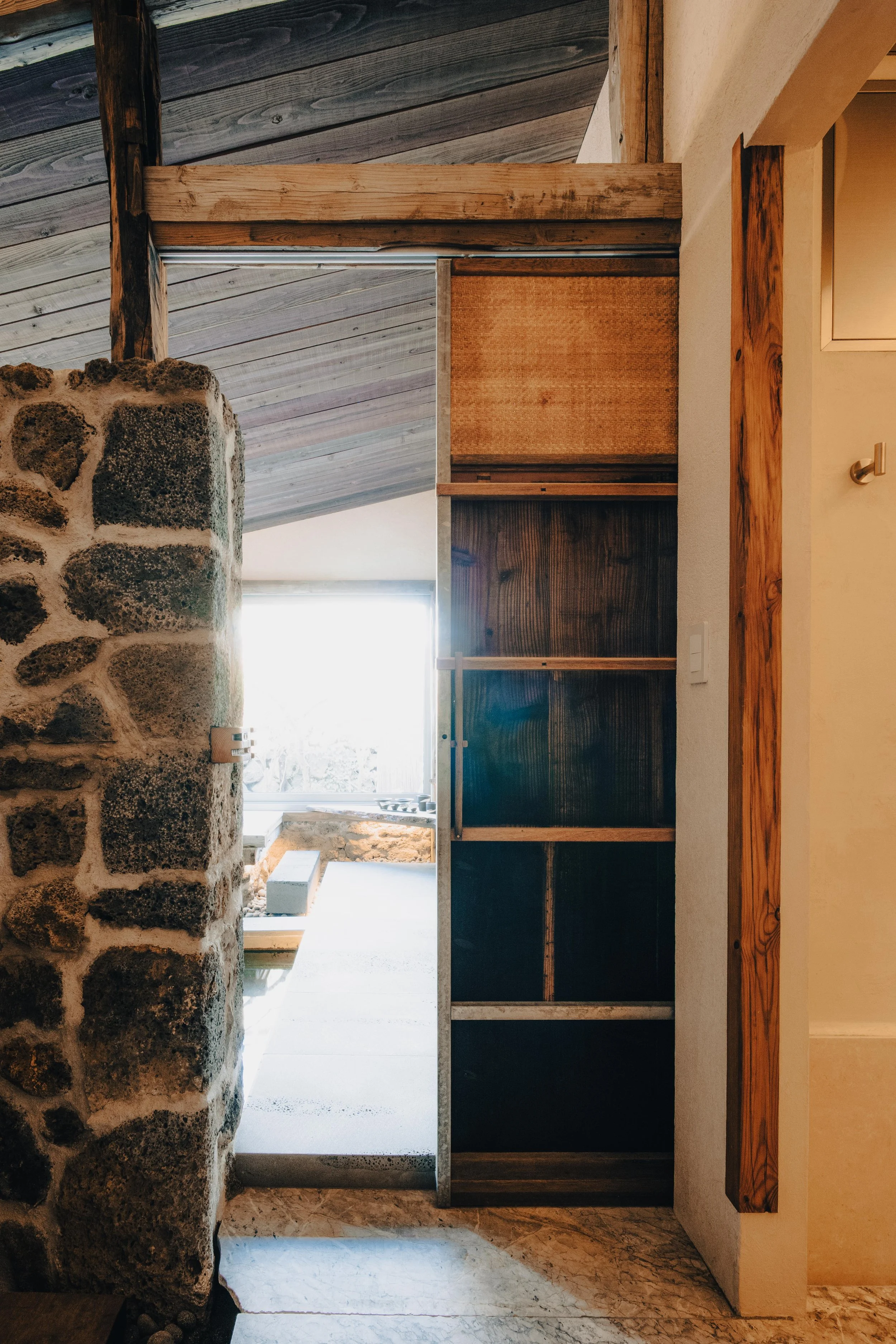NUUT Aewol 눗애월 Interior view of a room with a stone wall, wooden beams, and a partially open sliding door with wooden shelves behind it. Natural light coming through a window in the background.