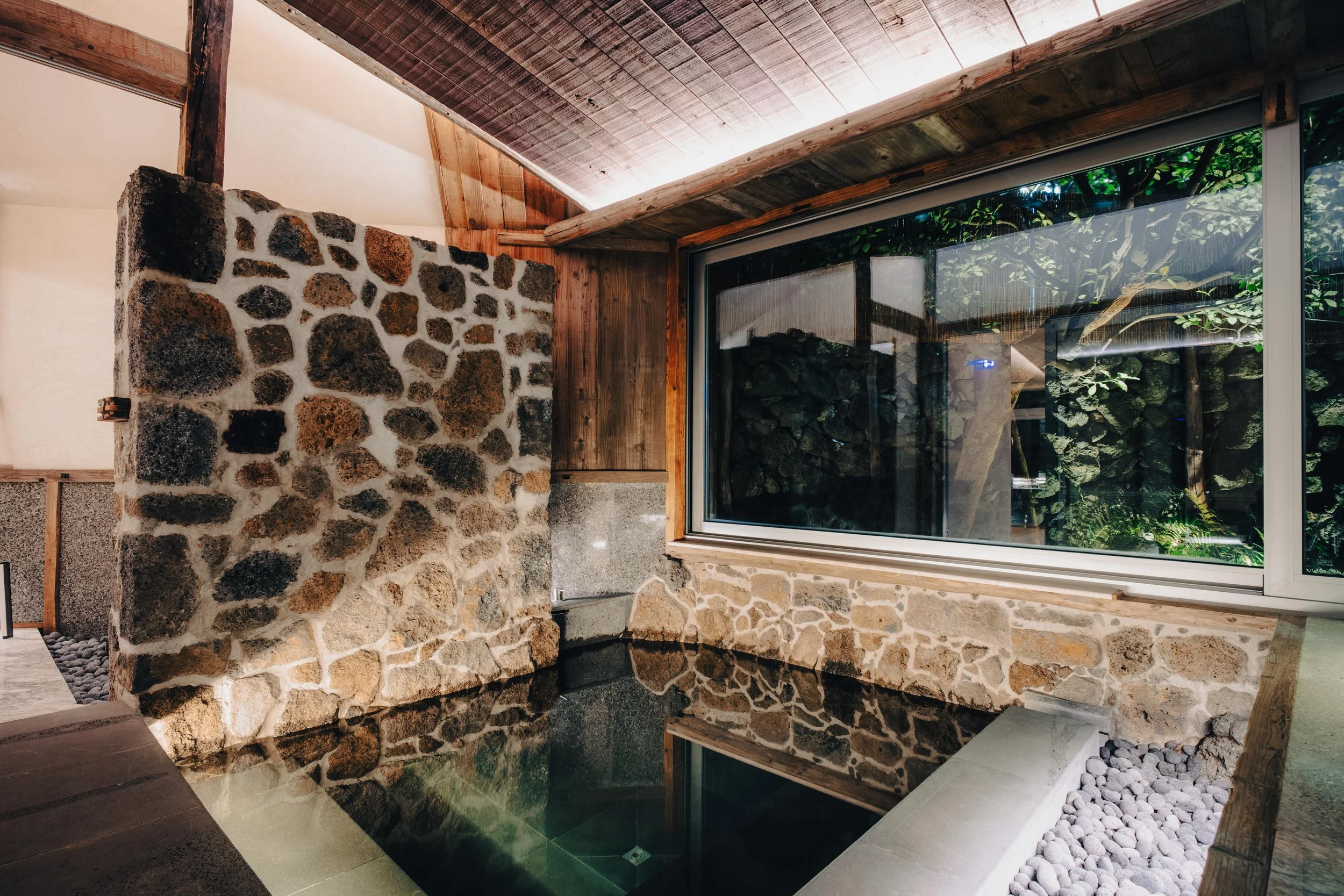 NUUT Aewol 눗애월 Indoor hot spring with a stone wall, large window showing greenery outside, and traditional wooden interior design.