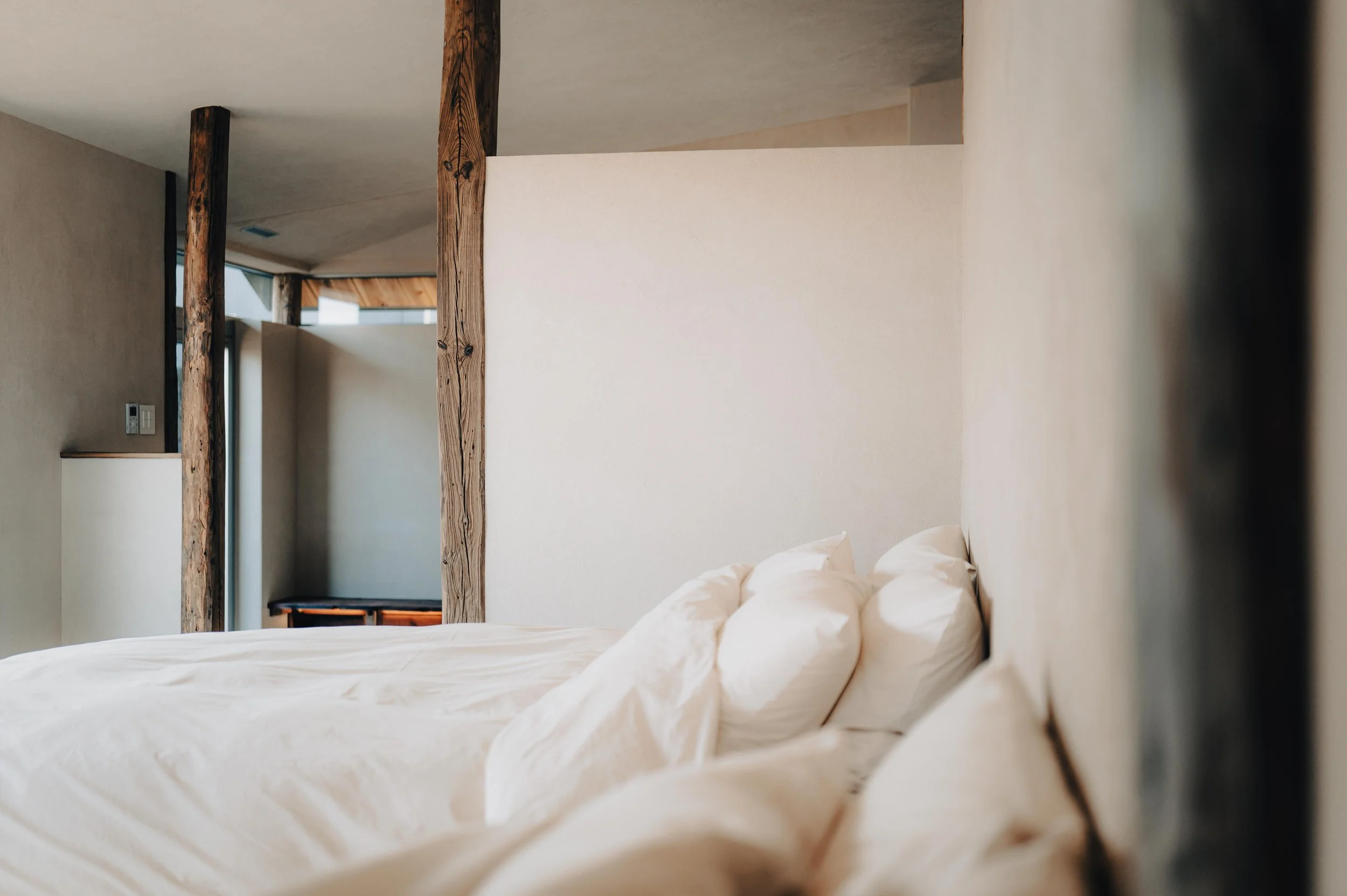 NUUT Aewol 눗애월 A neatly made bed with white linens and multiple pillows in a minimalistic bedroom with beige walls, wooden beams, and a glimpse of a window.