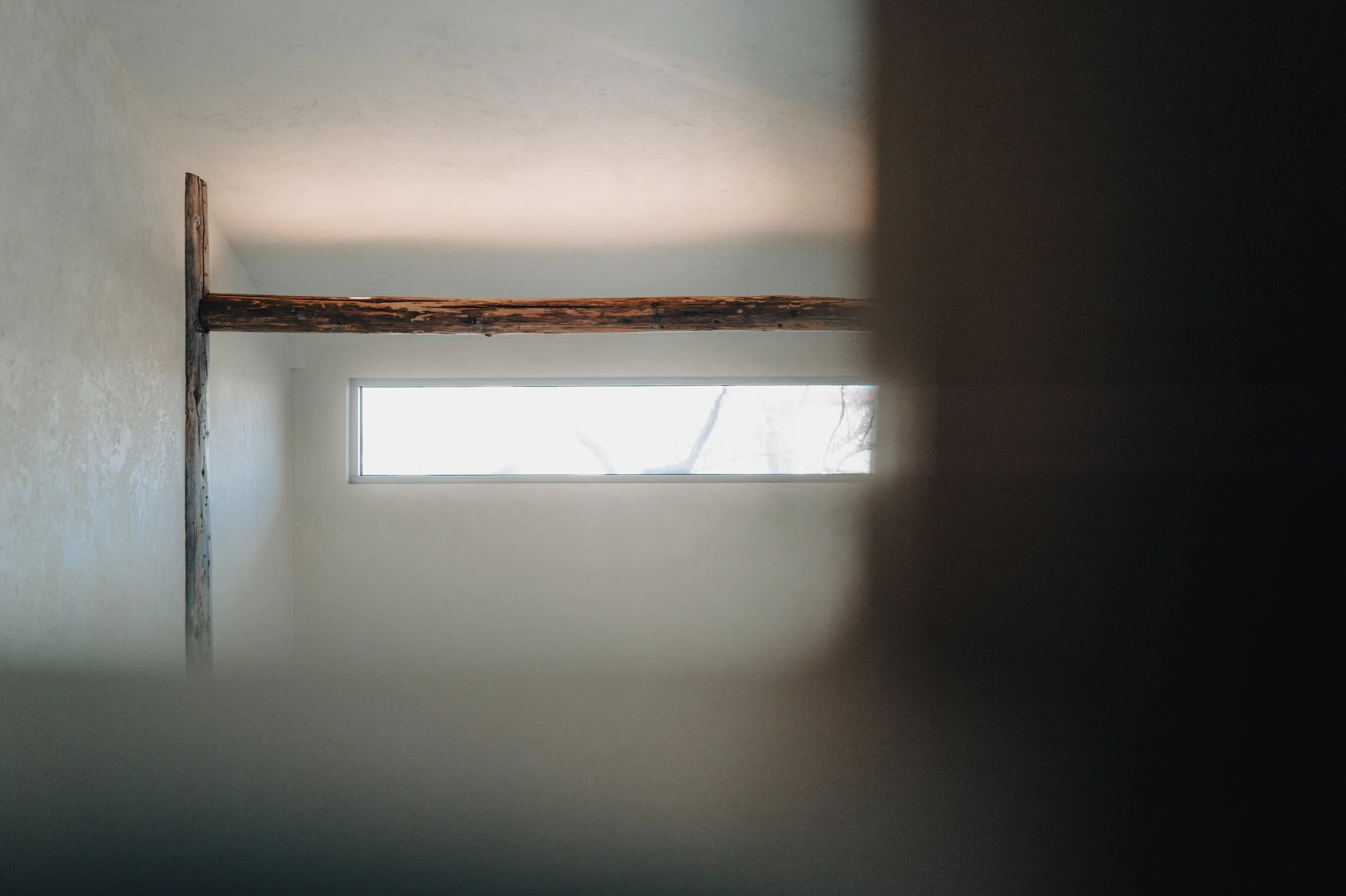 NUUT Aewol 눗애월 Interior view of a room with white walls, a horizontal window, and a rustic wooden beam crossing the ceiling.