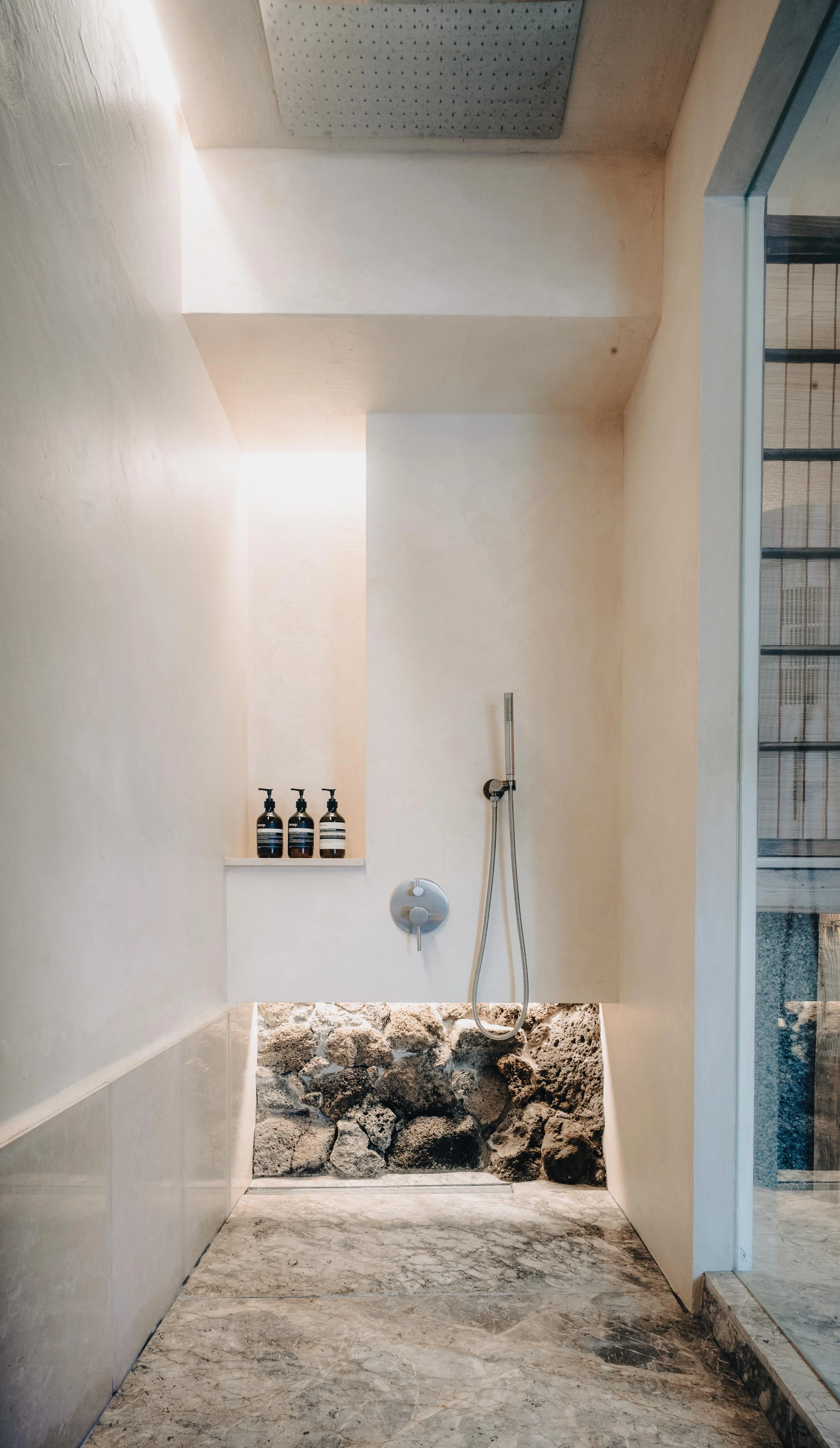NUUT Aewol 눗애월 Minimalist shower area with natural stone and rocks, with three bottles of soap or shampoo on a ledge, and a handheld showerhead mounted on the wall.