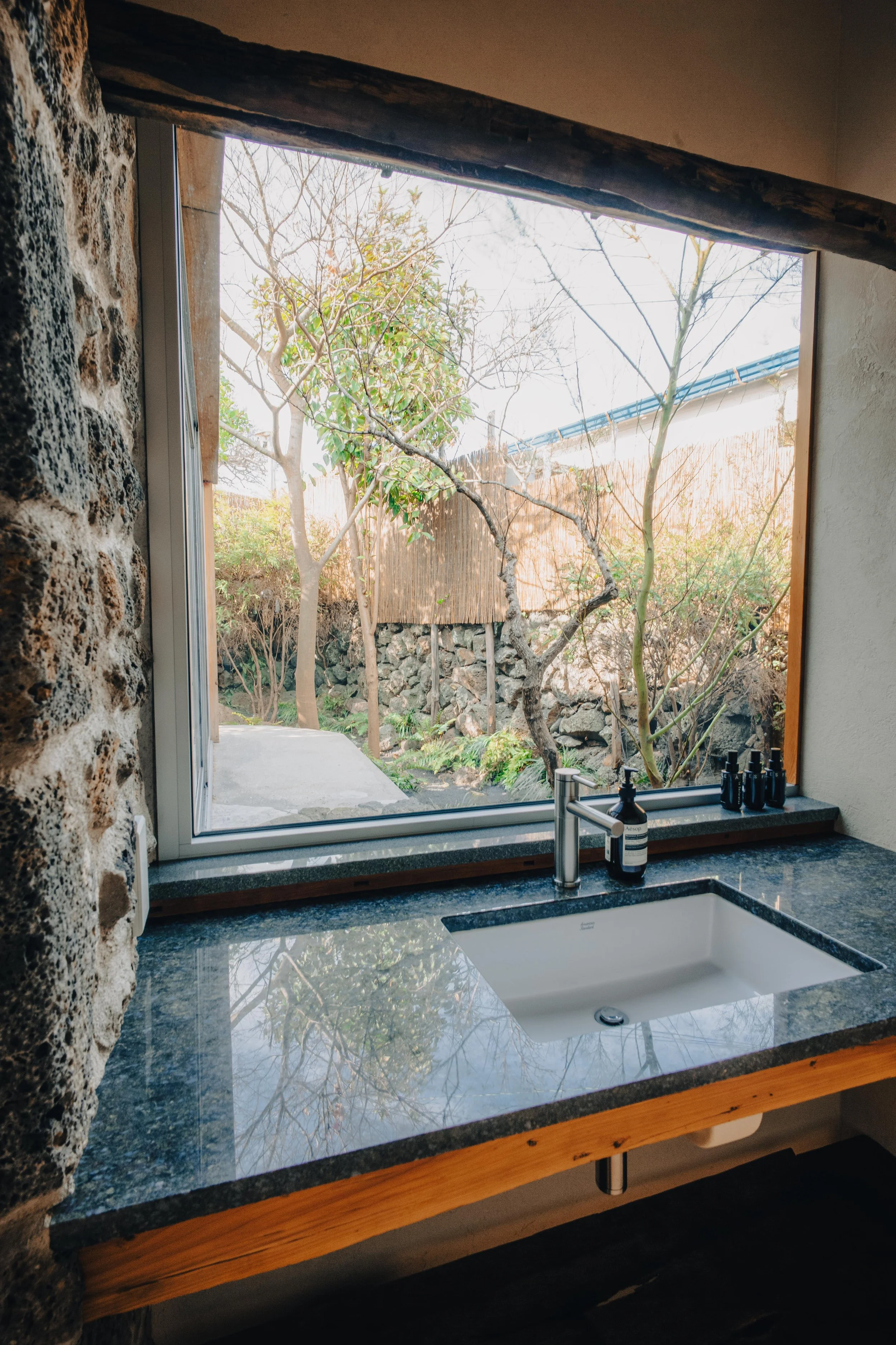 NUUT Aewol 눗애월 View of a window in a bathroom with a stone wall and a granite countertop with a white sink, and a garden with trees and rocks outside.