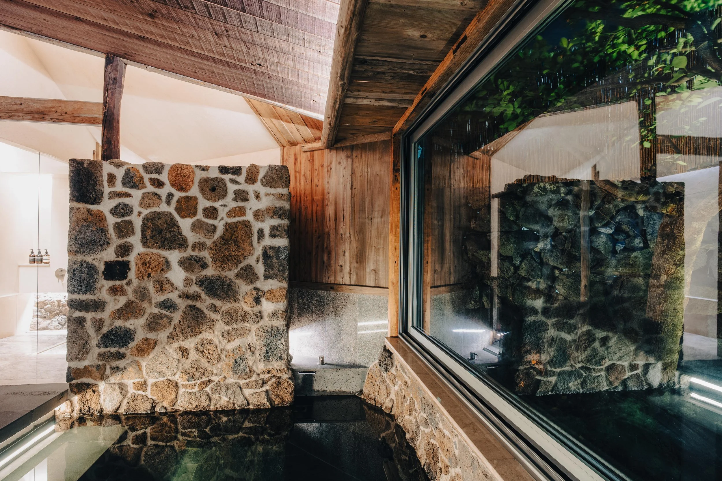 NUUT Aewol 눗애월 Interior view of a room featuring a stone wall, a large window reflecting a stone structure outside, and wooden ceiling and wall panels.
