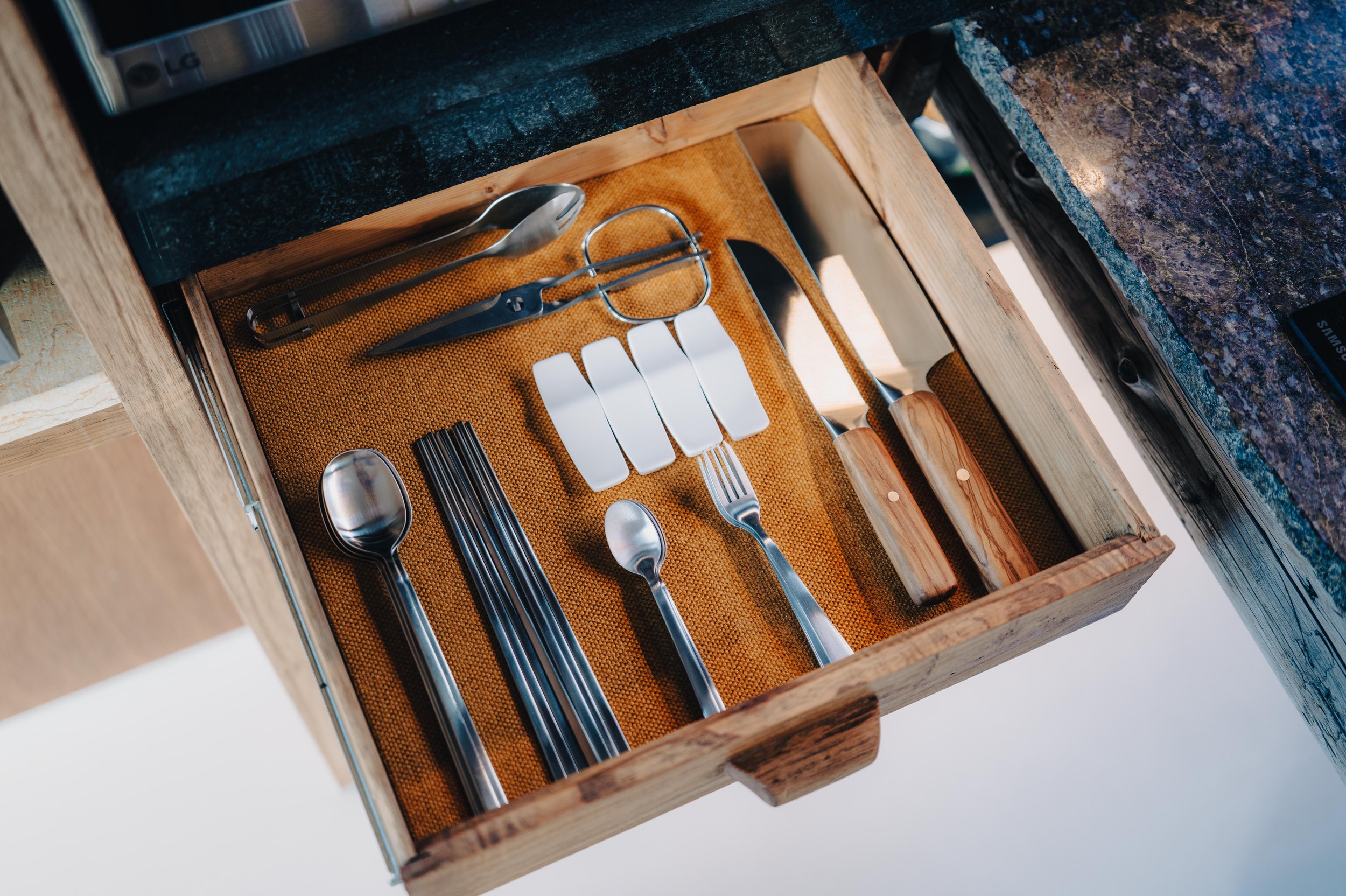 NUUT Aewol 눗애월 Kitchen drawer containing various kitchen utensils including a spoon, scissors, tongs, butter knife, multiple metal forks, a small spoon, a potato peeler, and some white tags, all organized on a brown textured liner.