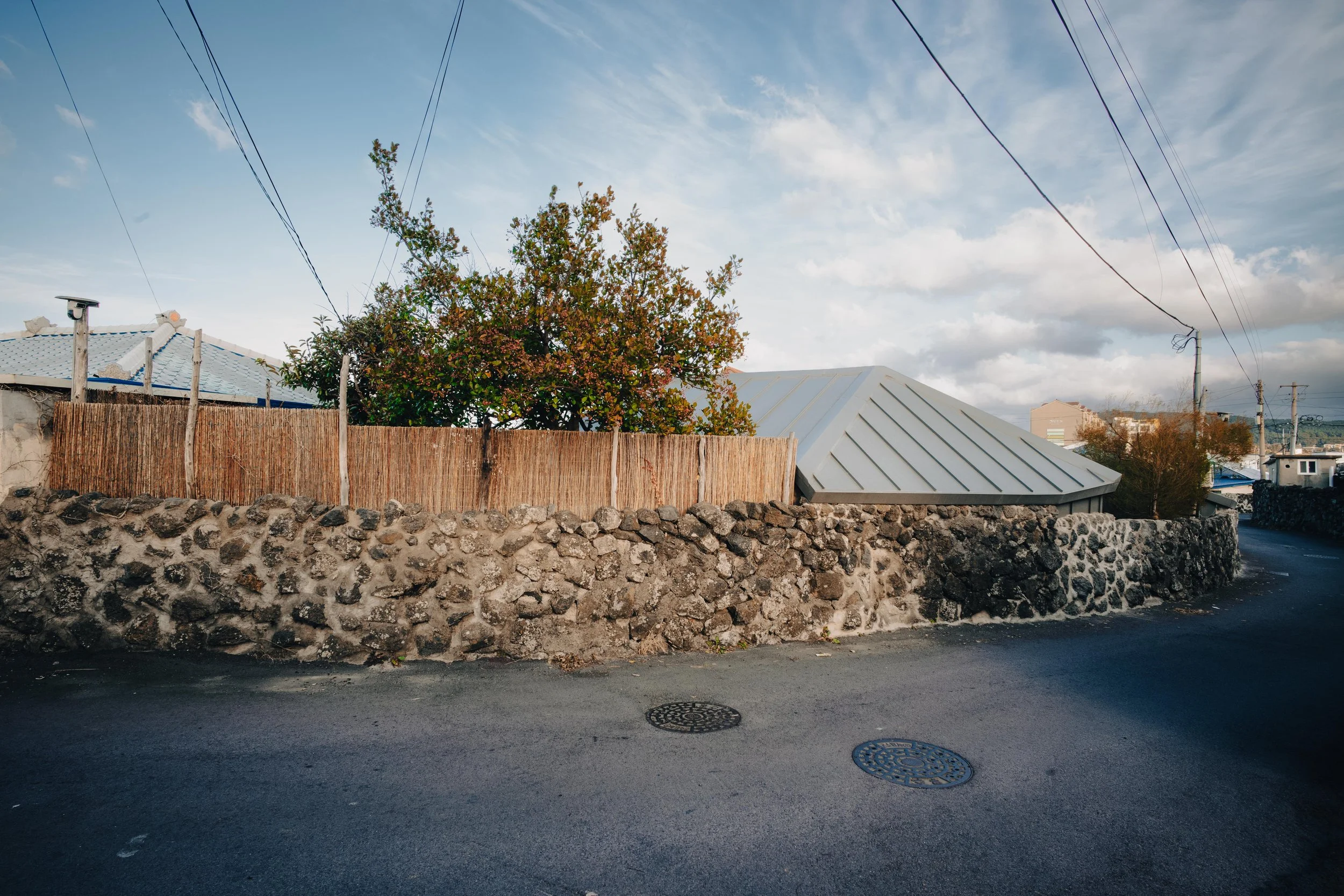 NUUT Aewol 눗애월 A residential street scene showing a stone wall, wooden fence, trees, a house with a metal roof, utility poles, and a cloudy sky.