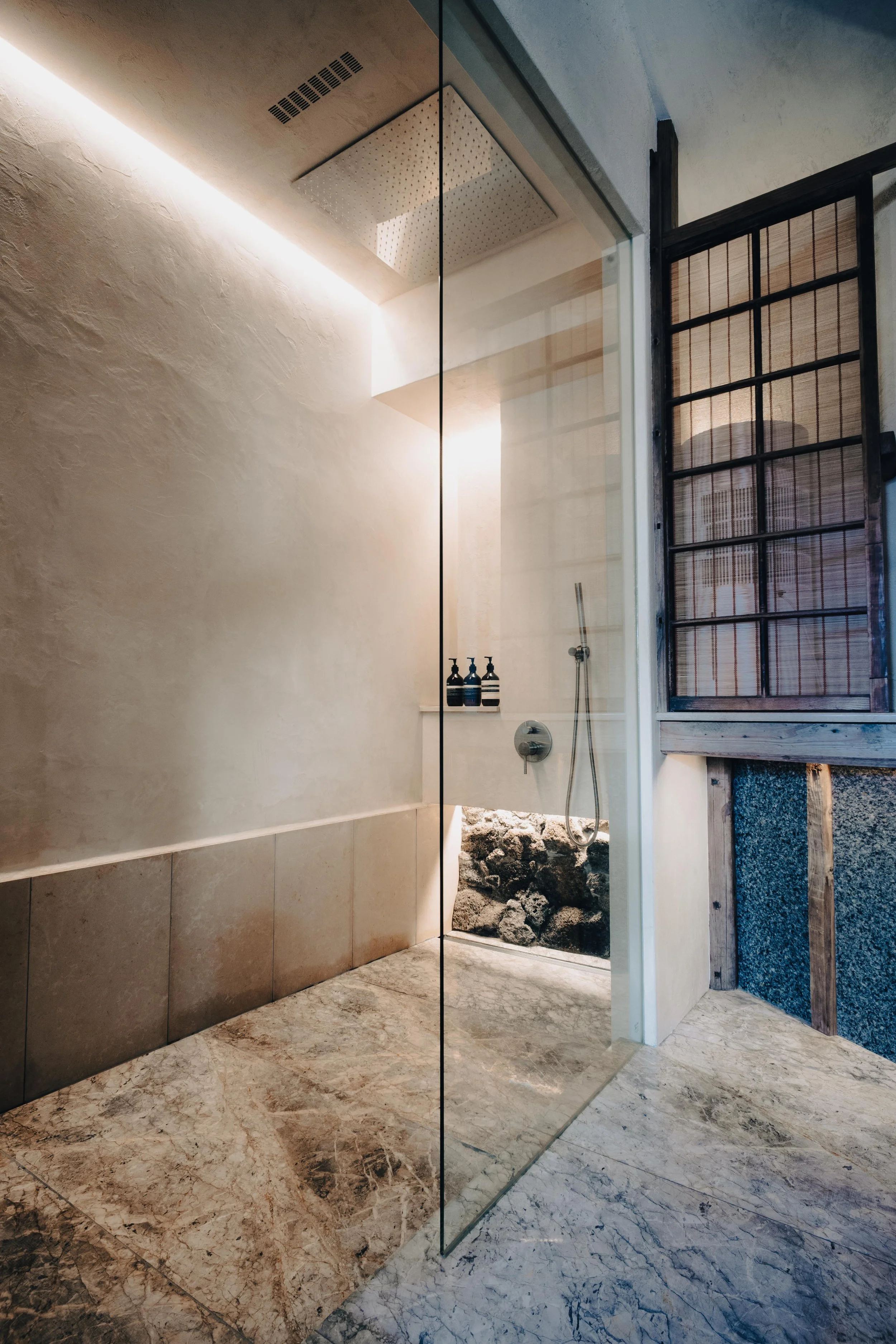NUUT Aewol 눗애월 Modern bathroom with a walk-in shower featuring a glass door, stone accent on the shower floor, and natural light coming from a window with wooden framing.