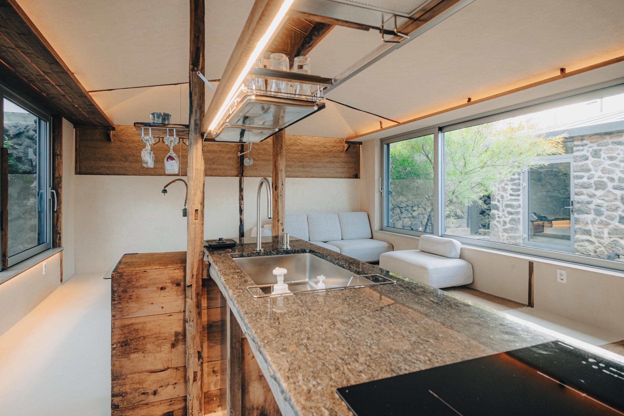 NUUT Aewol 눗애월 Modern kitchen with granite countertop, wooden cabinetry, and large windows overlooking a stone wall and greenery.