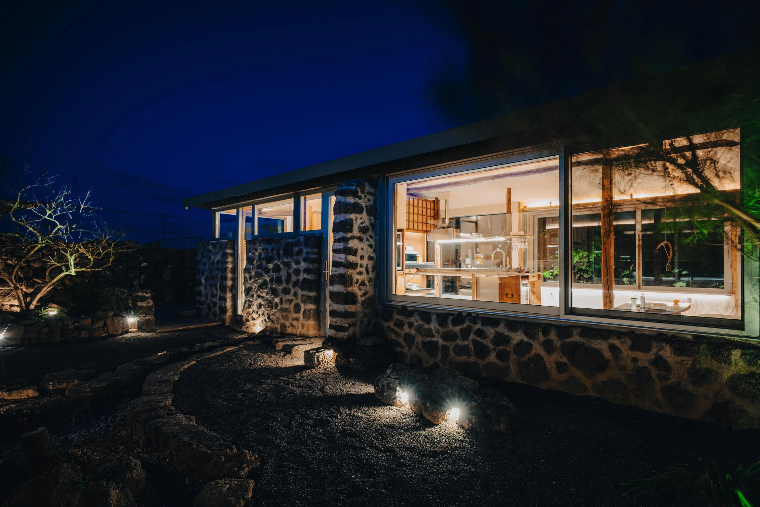 NUUT Aewol 눗애월 Night view of a modern house with large floor-to-ceiling windows revealing a well-lit interior, stone wall exterior, and a small illuminated outdoor garden with rocks and a tree.