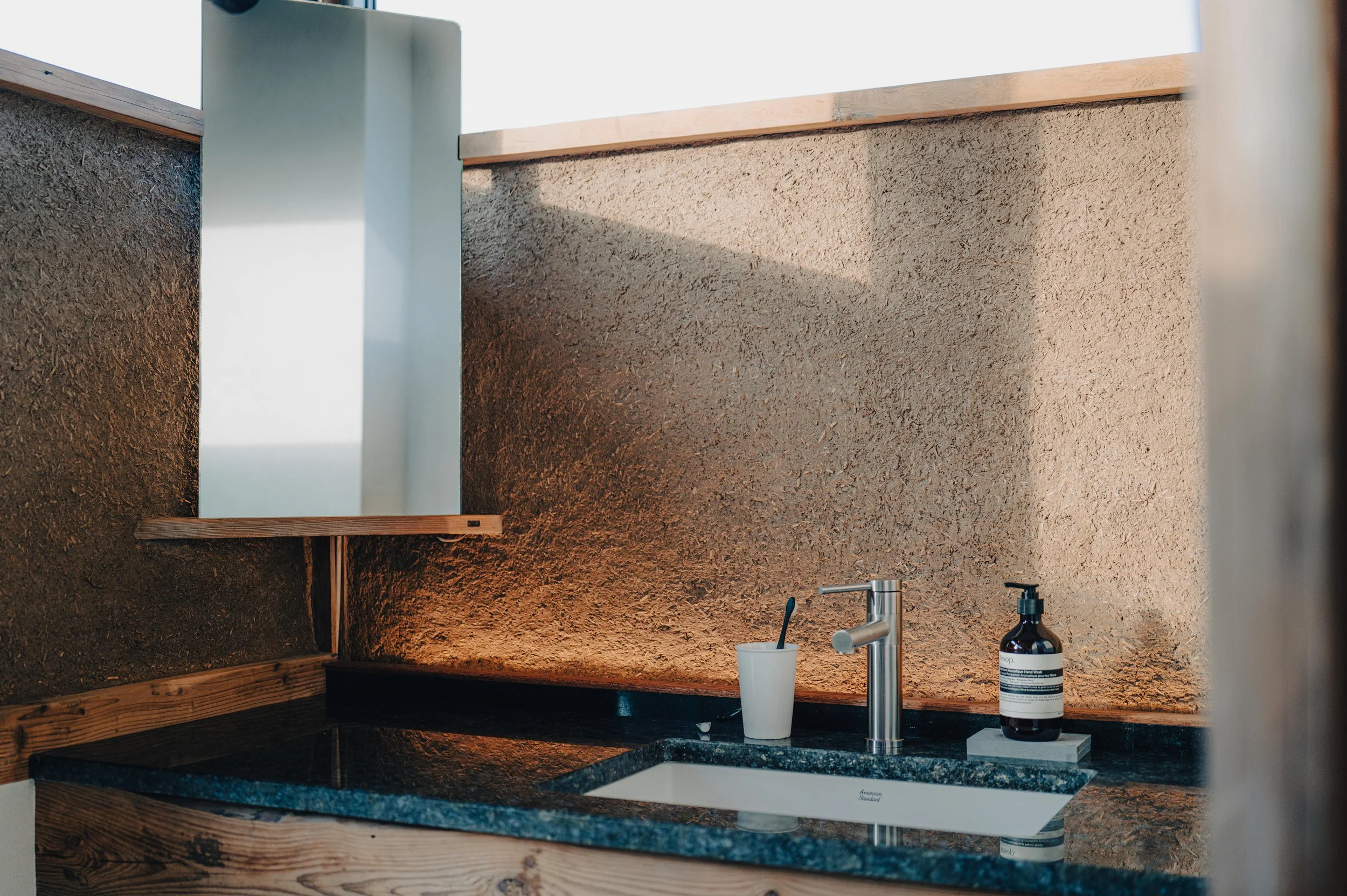 NUUT Aewol 눗애월 Close-up of a bathroom or kitchen sink area with a modern stainless steel faucet, a white cup with a toothbrush, a soap dispenser, a black granite countertop, a round mirror, and textured beige walls.