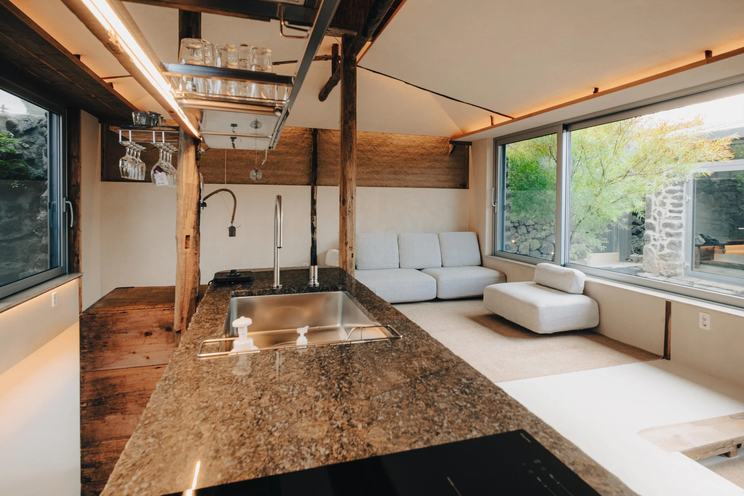 NUUT Aewol 눗애월 Open-concept living room and kitchen with large windows, light-colored sofa, stone accents, and wooden beams, overlooking a garden with trees.