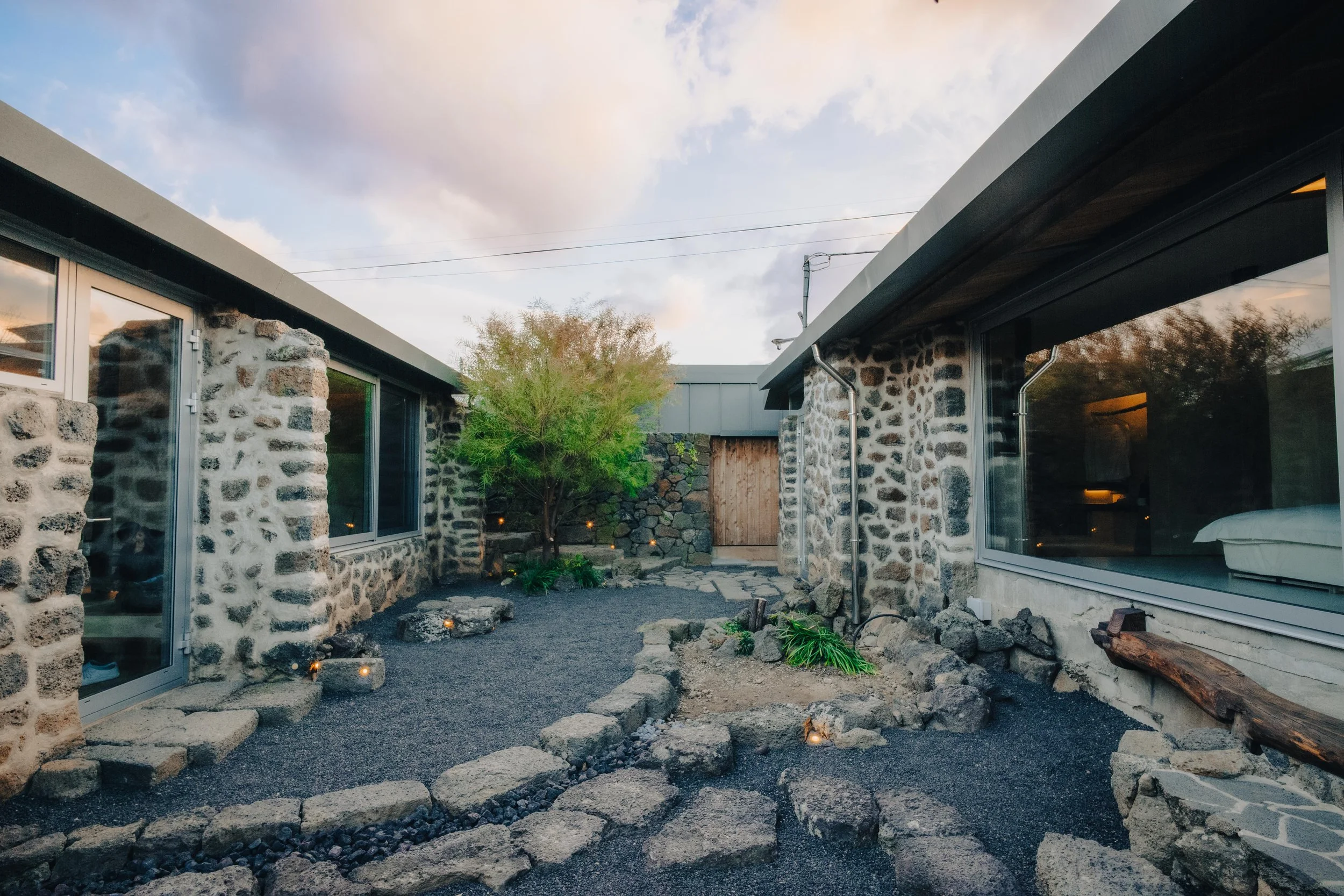 NUUT Aewol 눗애월 A modern residential courtyard with stone walls, large windows, potted plants, and a pathway made of rocks, illuminated by small ground lights at dusk.