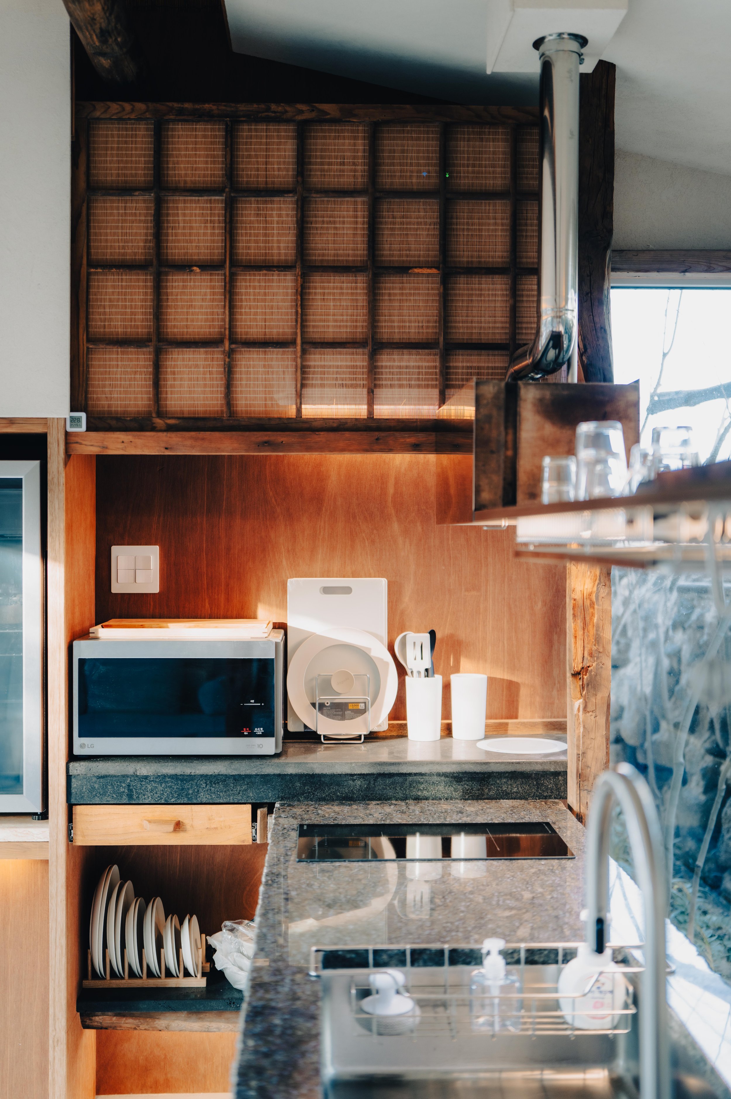 NUUT Aewol 눗애월 Kitchen countertop with microwave, rice cooker, paper towels, utensil holder, and electric stovetop. Wooden shelves with plates and glasses above and below the counter. Window on the right.