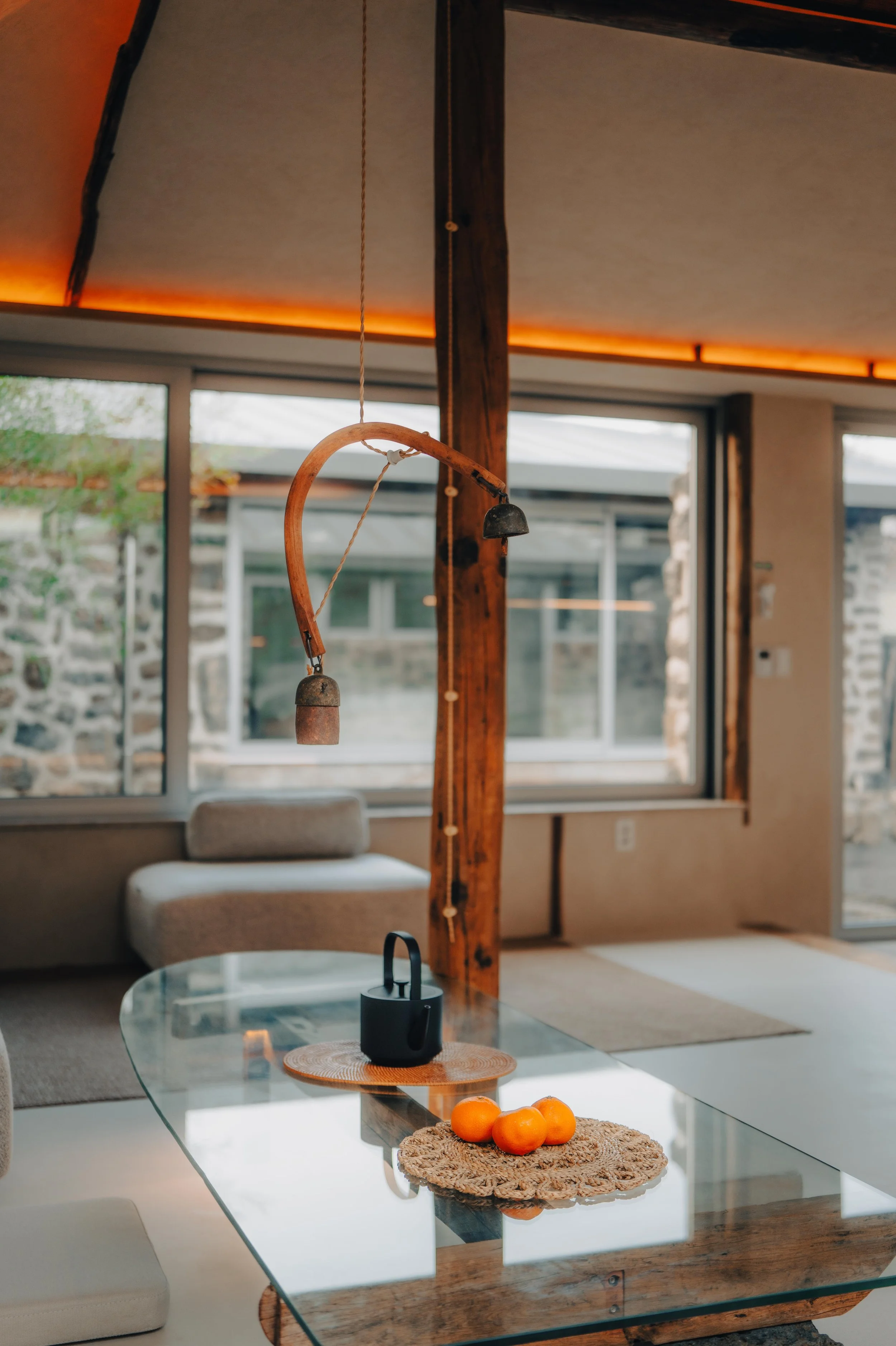 NUUT Aewol 눗애월 Interior of a modern living room with a glass-top table, a black teapot, and a woven placemat with three oranges. Large windows with views of stone walls and greenery outside. A wooden beam with hanging decorative elements.
