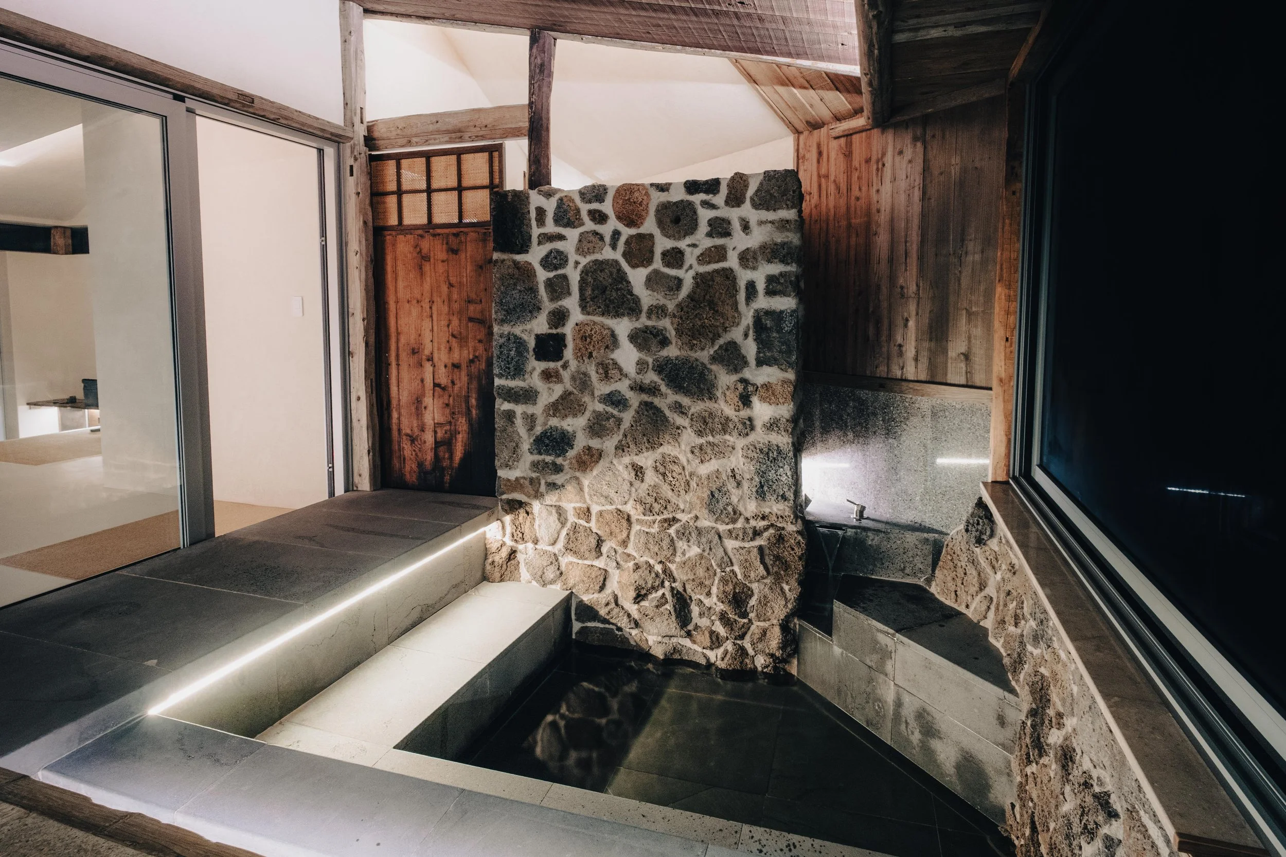 NUUT Aewol 눗애월 Indoor space featuring a stone wall, a small fountain or water feature, wooden walls and ceiling, a glass window, and sliding glass doors leading to another room.