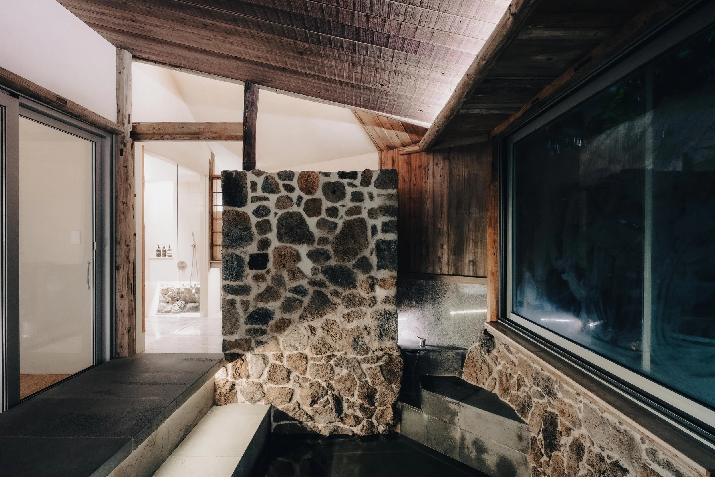 NUUT Aewol 눗애월 Indoor space with stone wall, large window, wooden ceiling and walls, stone bench, and a glimpse of a bathroom in the background.
