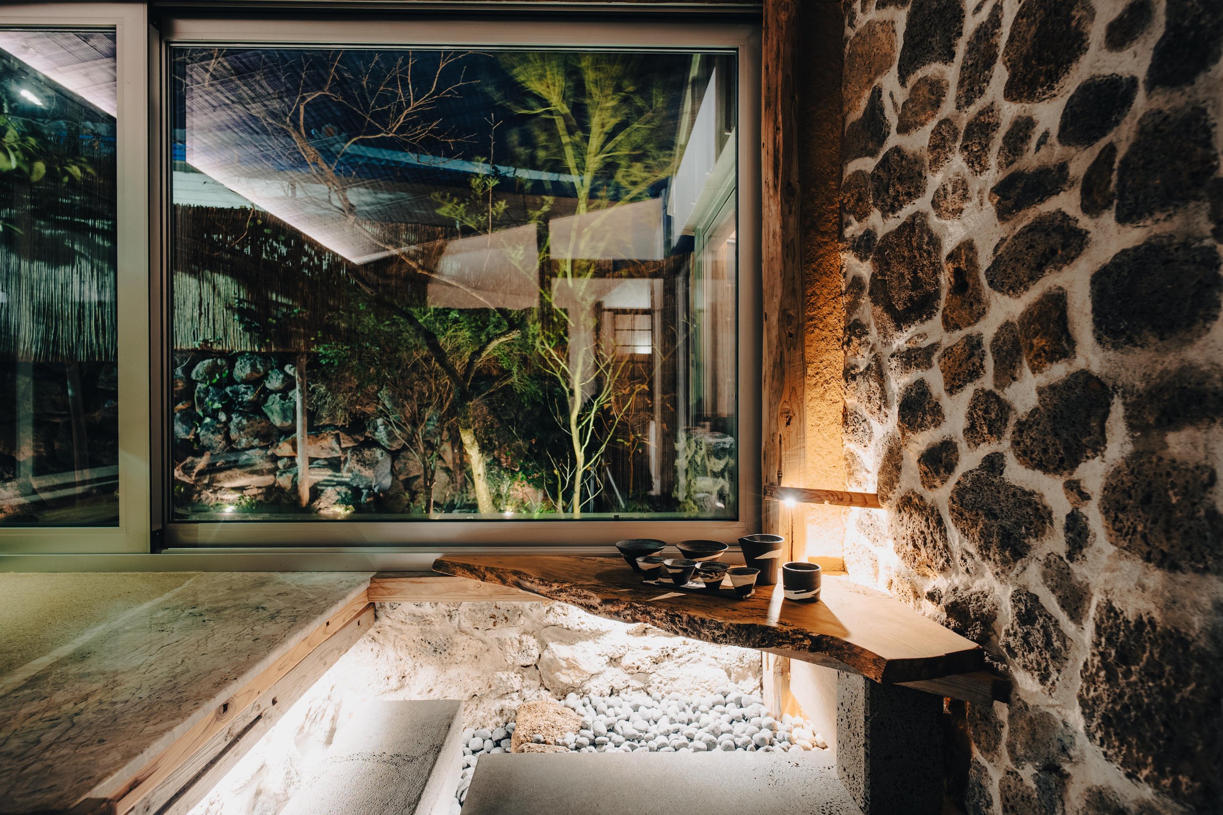 NUUT Aewol 눗애월 Interior view of a rustic stone wall near a window with a wooden ledge holding black and white ceramic bowls, with a garden visible outside through the window.