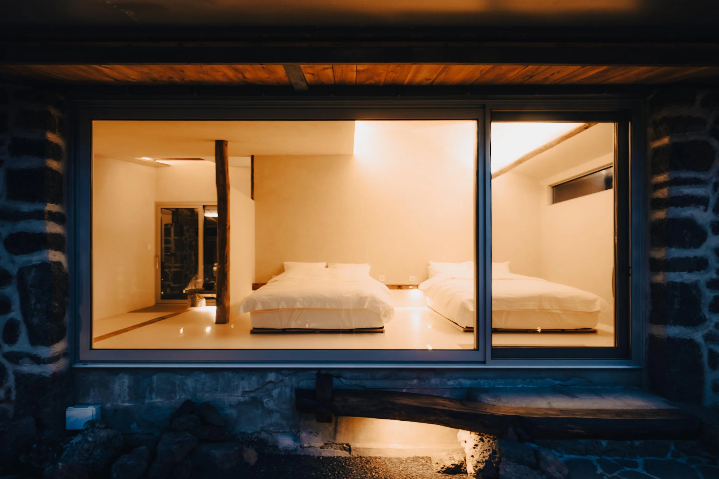 NUUT Aewol 눗애월 View through a large window of a modern bedroom with two beds, wooden accents, and soft lighting, seen from outside at night.
