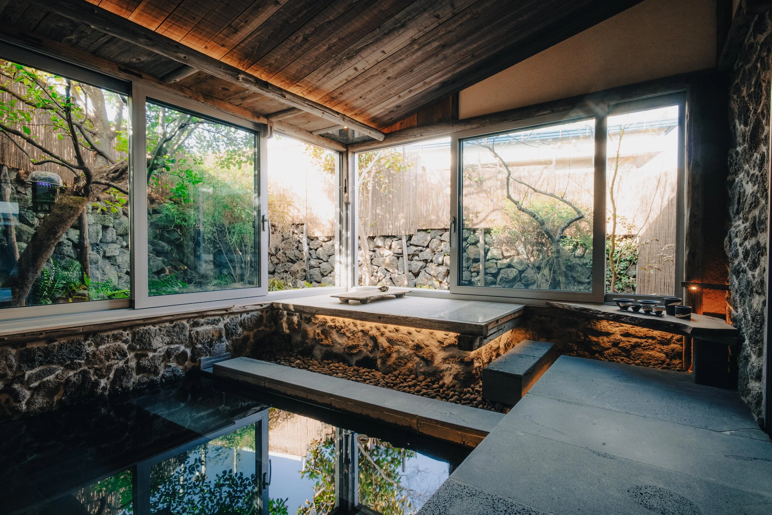 Interior of a traditional Japanese-style room with large sliding glass doors, stone walls, a small indoor pond, and a garden view outside.