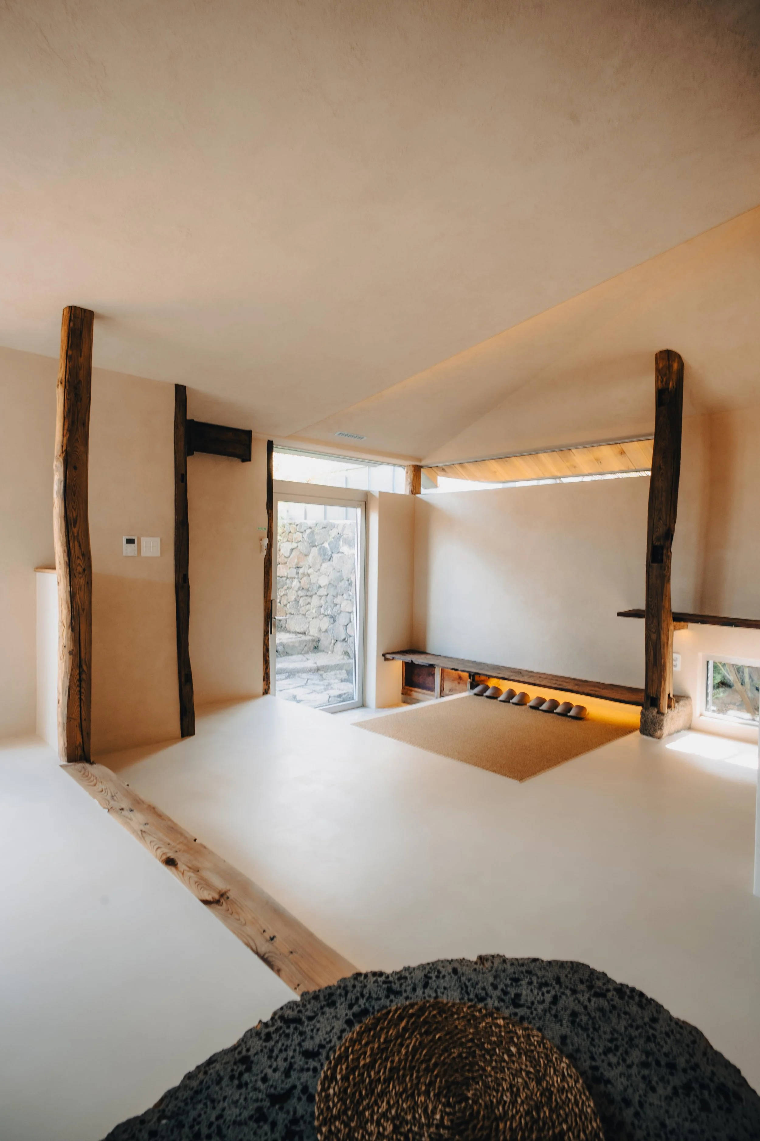 NUUT Aewol 눗애월 Interior view of a modern room with beige walls, wooden beams, a glass door, and natural light.