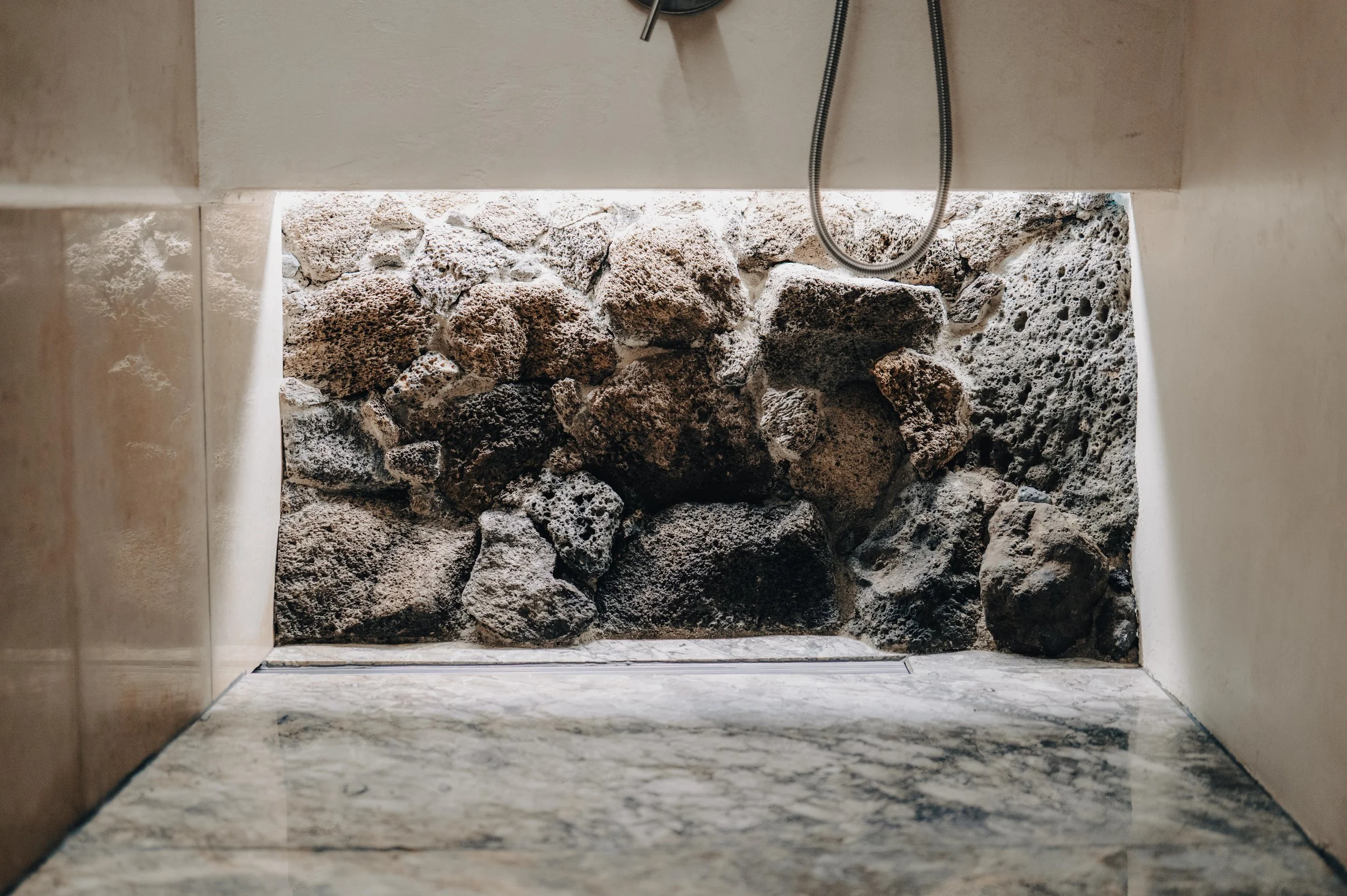 NUUT Aewol 눗애월 Close-up of a shower with a stone wall, marble floor, and a shower head with hose.