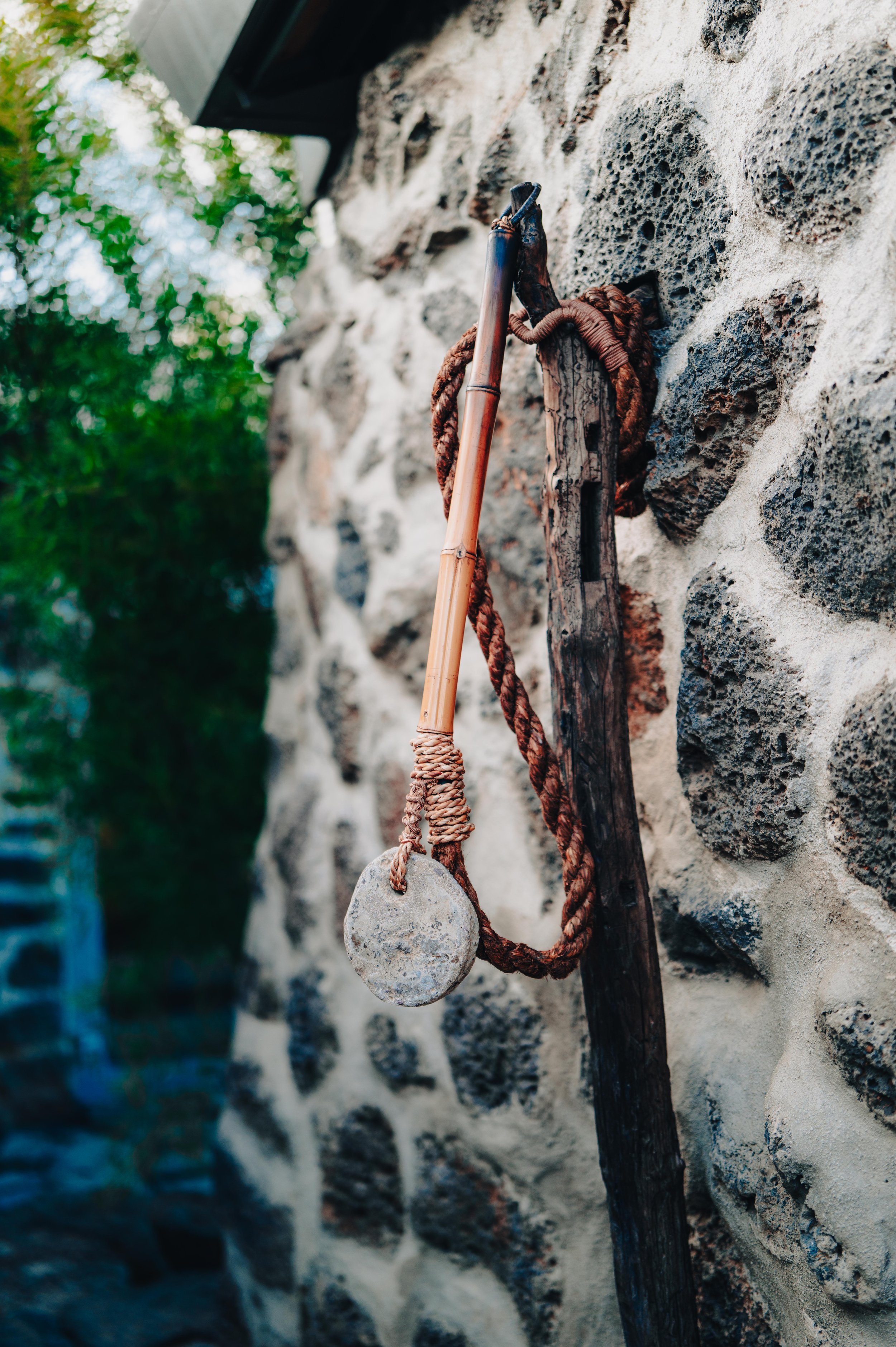 NUUT Aewol 눗애월 An ancient rock climbing tool, possibly a pike or pick, mounted on a rustic stone wall with a thick rope and wooden handle hanging from it.