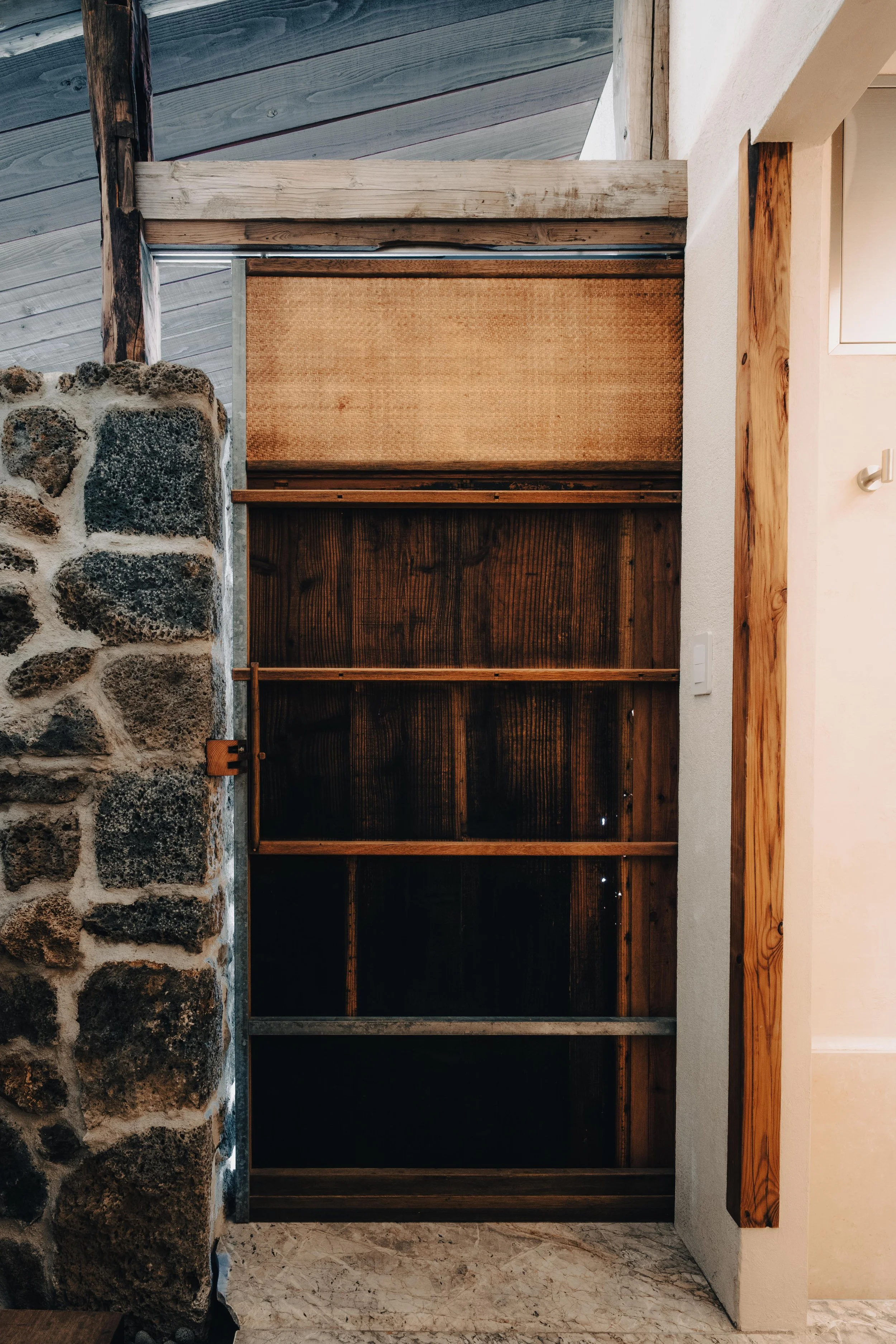 NUUT Aewol 눗애월 Interior view of a wooden and stone doorway with a sliding door, metal track, and exposed wooden beam ceiling.