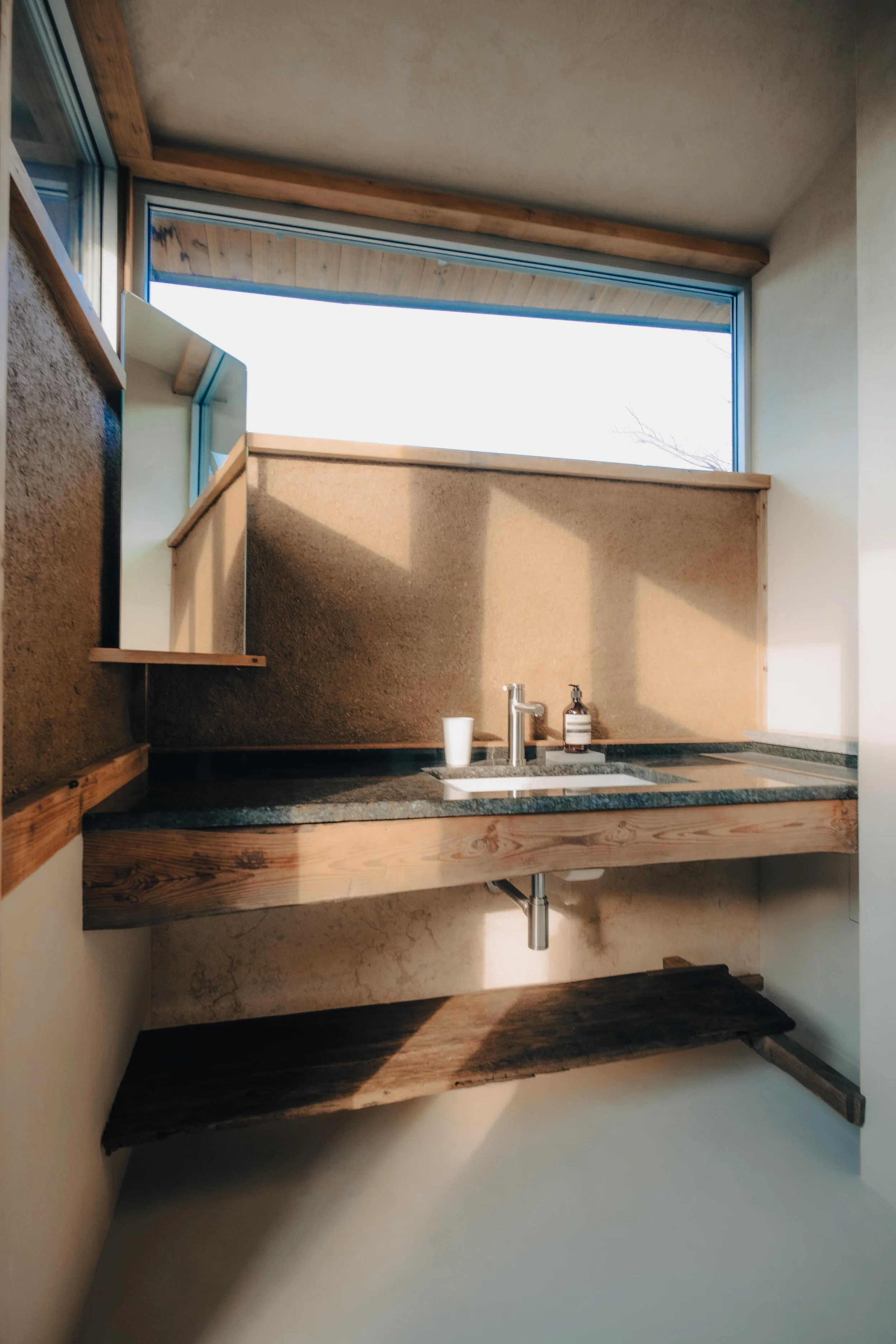 NUUT Aewol 눗애월 Interior view of a small kitchen with a single sink, a faucet, a soap dispenser, a cup, and wooden shelves under a large window.